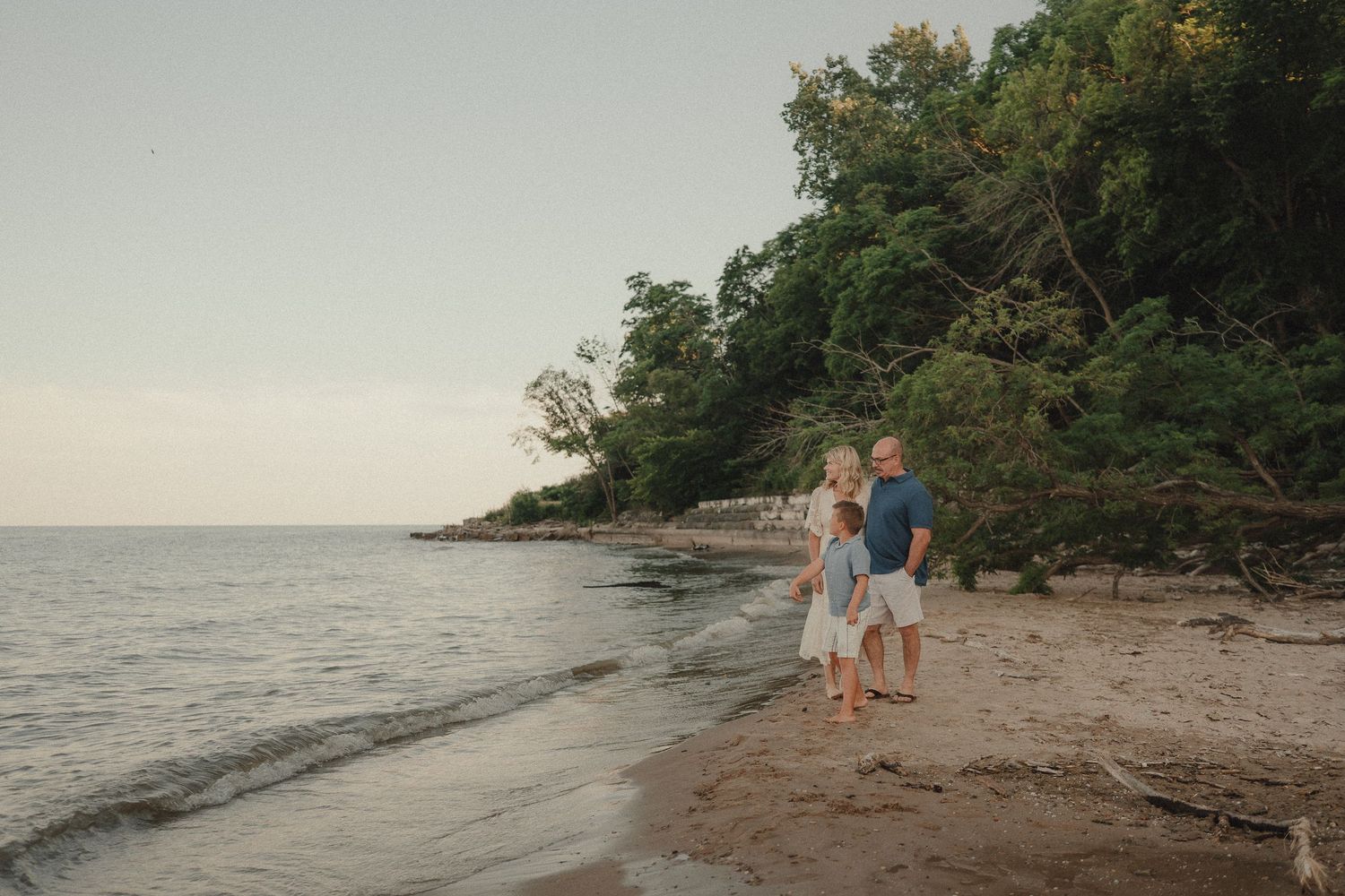 Family of three standing on a sandy beach at sunset, with lush green trees and calm water in the background.