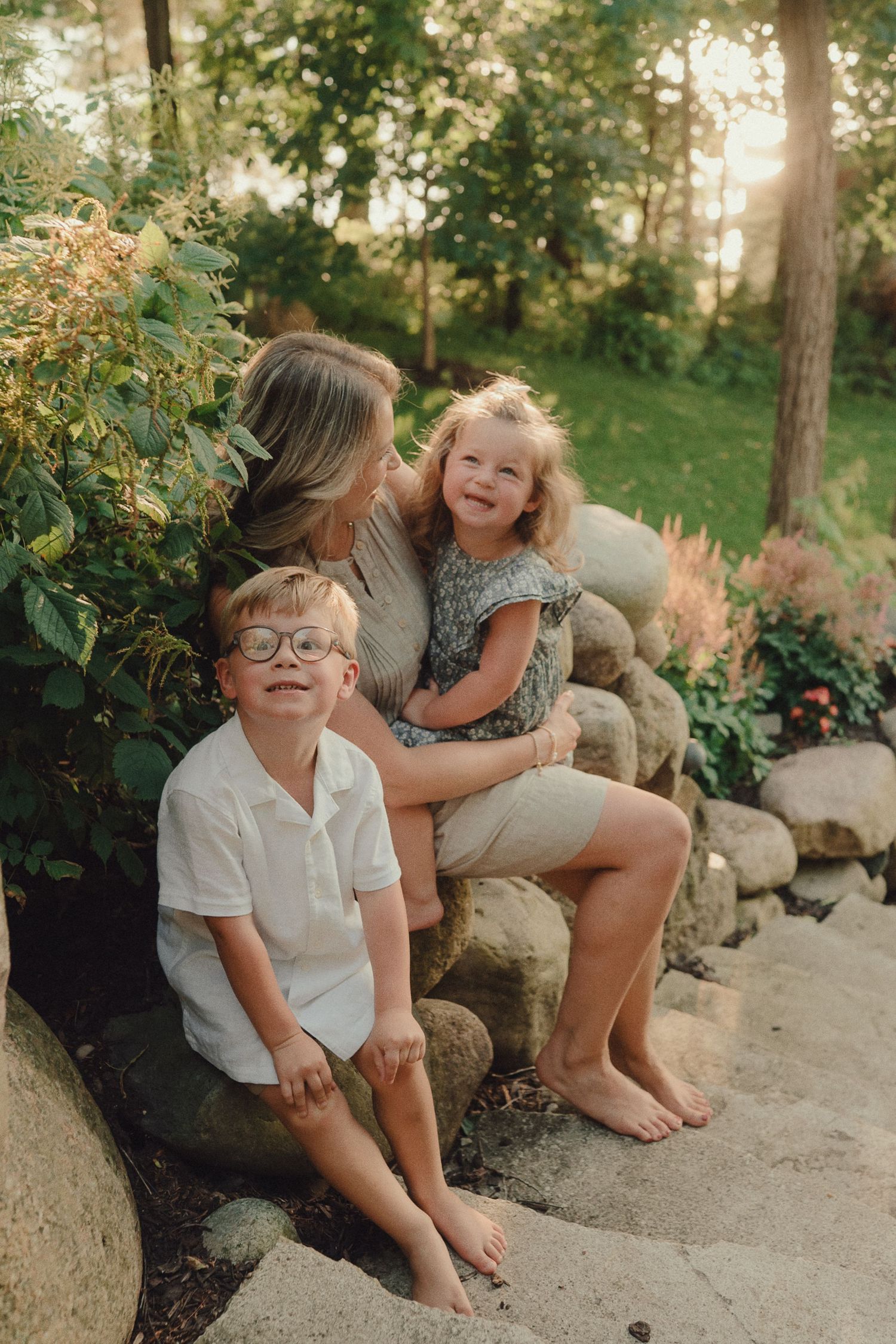 Mother sitting on stone steps with her two young children in a sunlit garden near a lake.