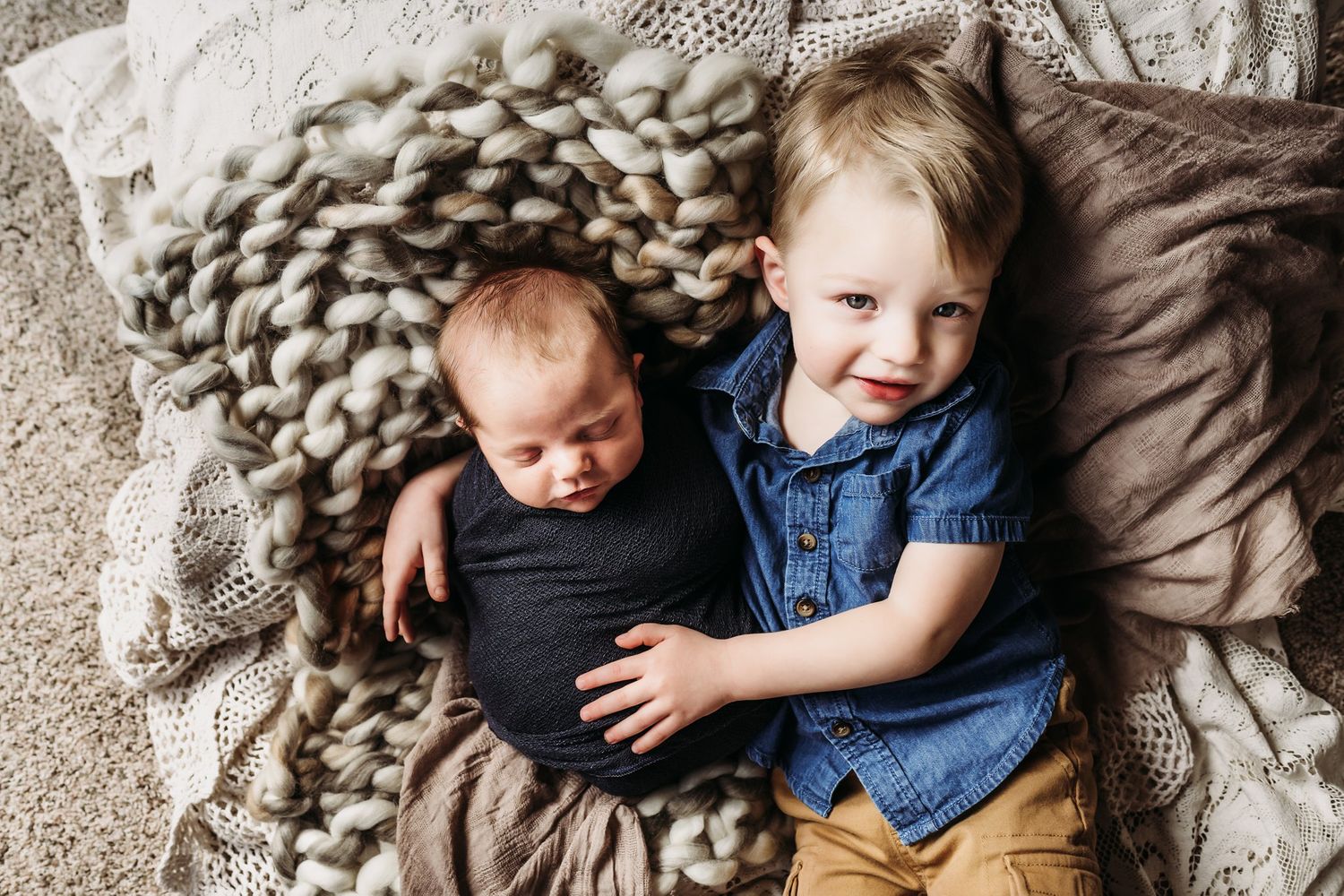 Two young children lying together on textured blanket with older sibling embracing baby.