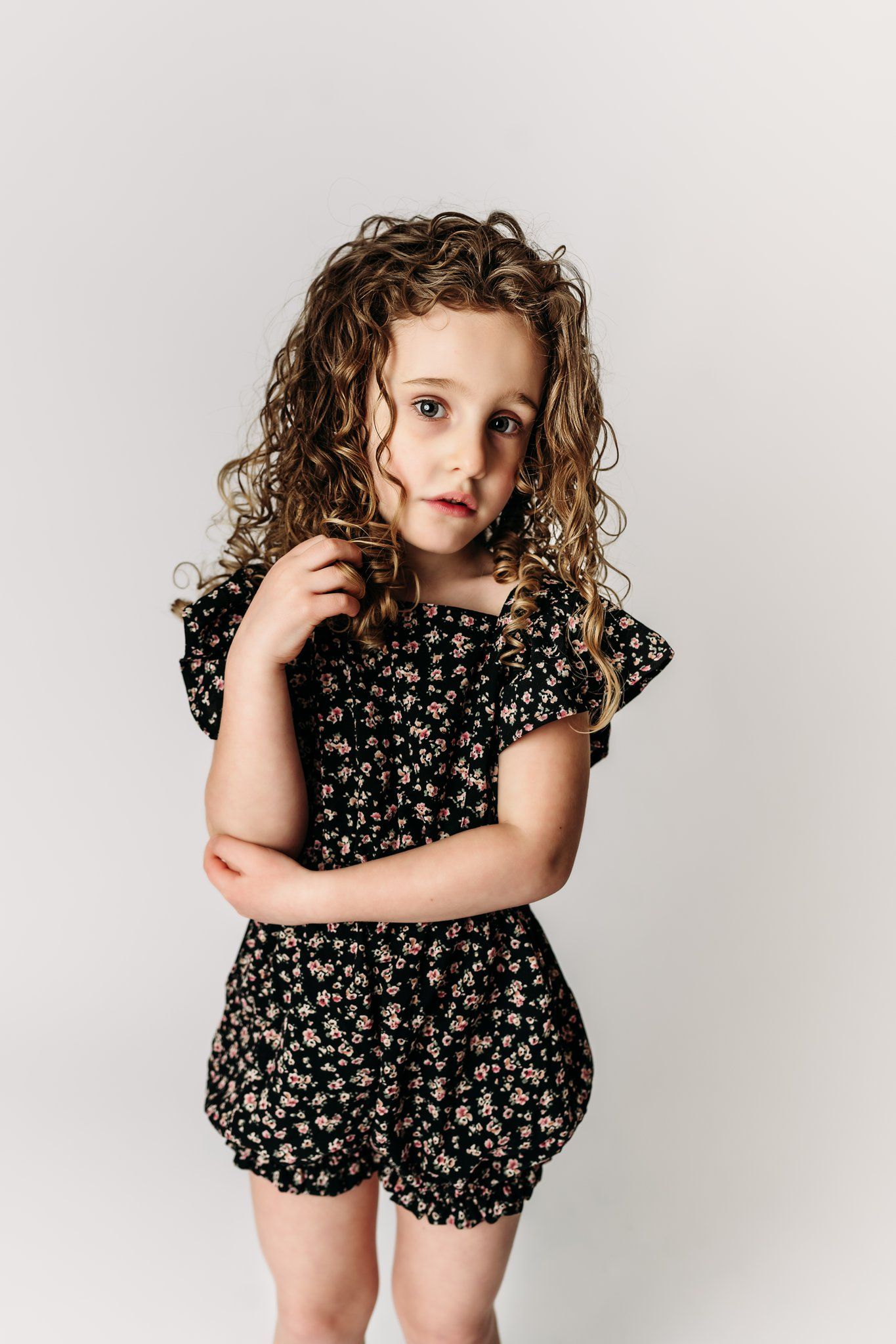 A child in a patterned romper stands shyly against white backdrop.