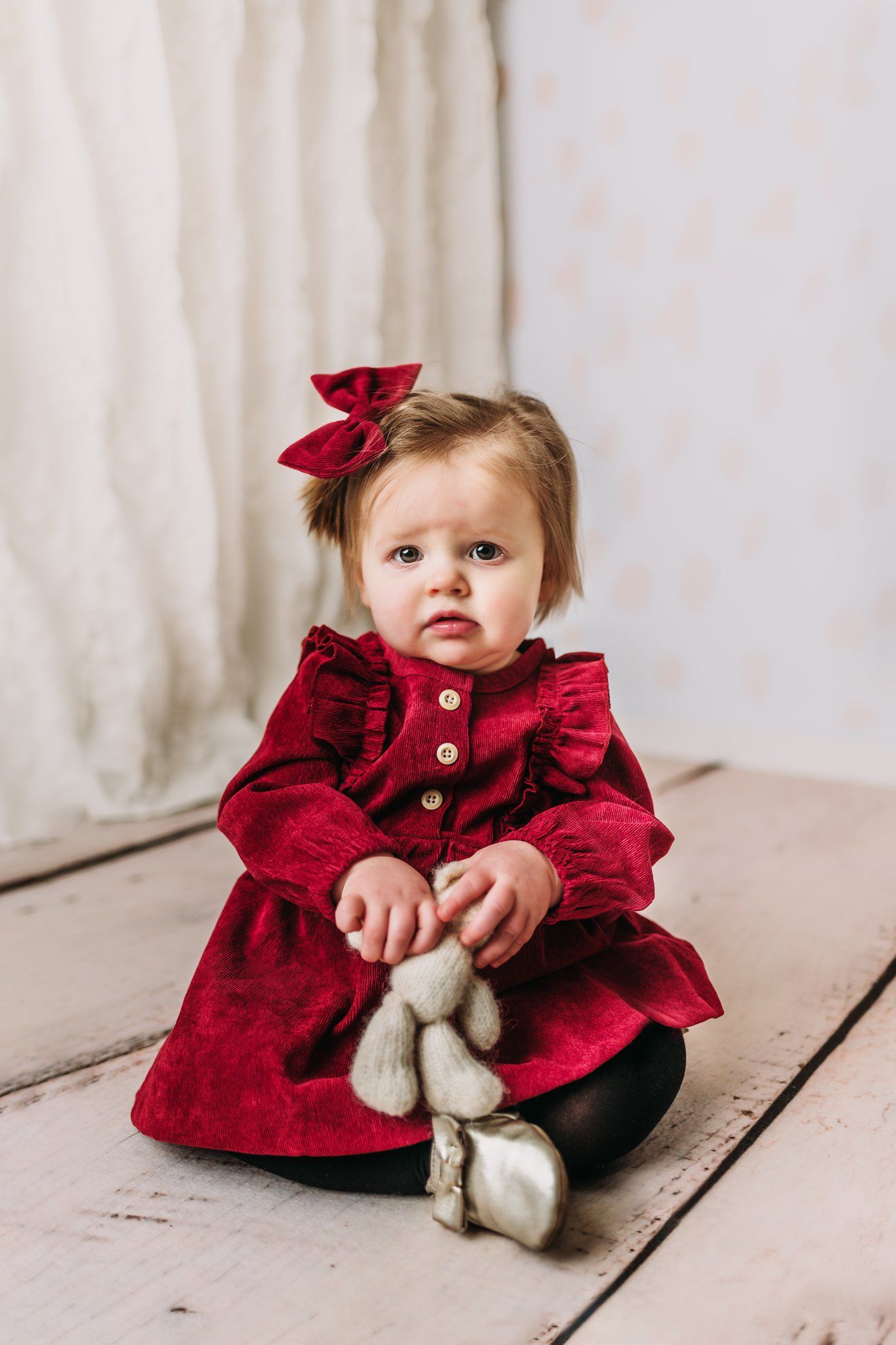 Baby in red velvet dress holds plush toy while sitting on wooden floor.