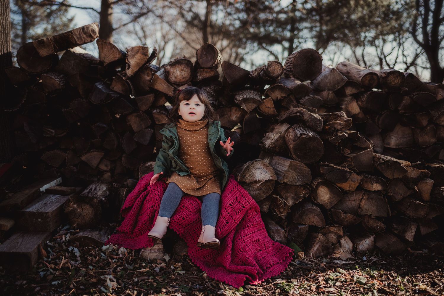 Child sitting joyfully on pink fabric against a woodpile background during sunset.
