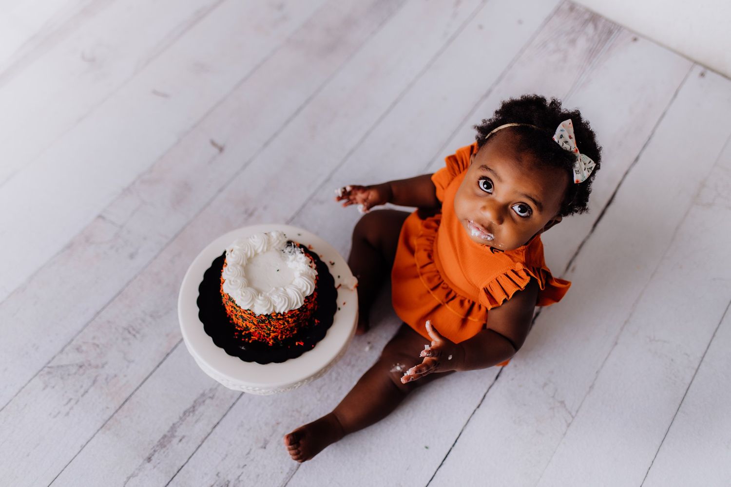 An overhead view of a small child in orange dress next to a decorated cake on the floor.