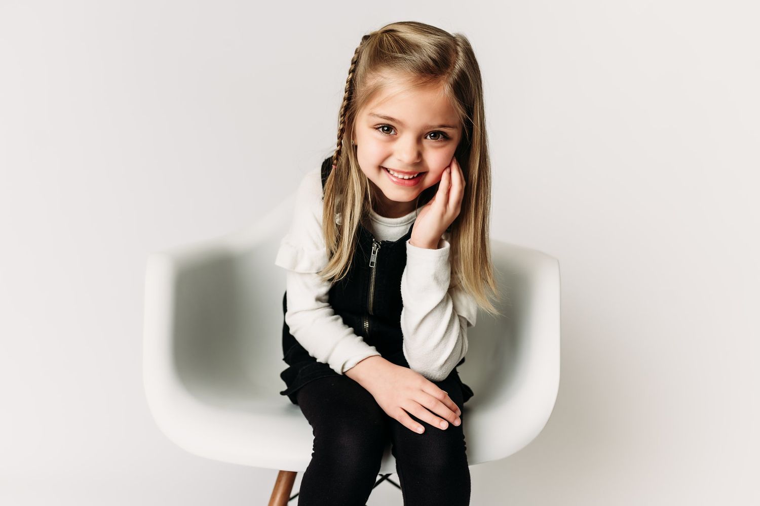 Child in white and black outfit poses playfully in modern chair setting.