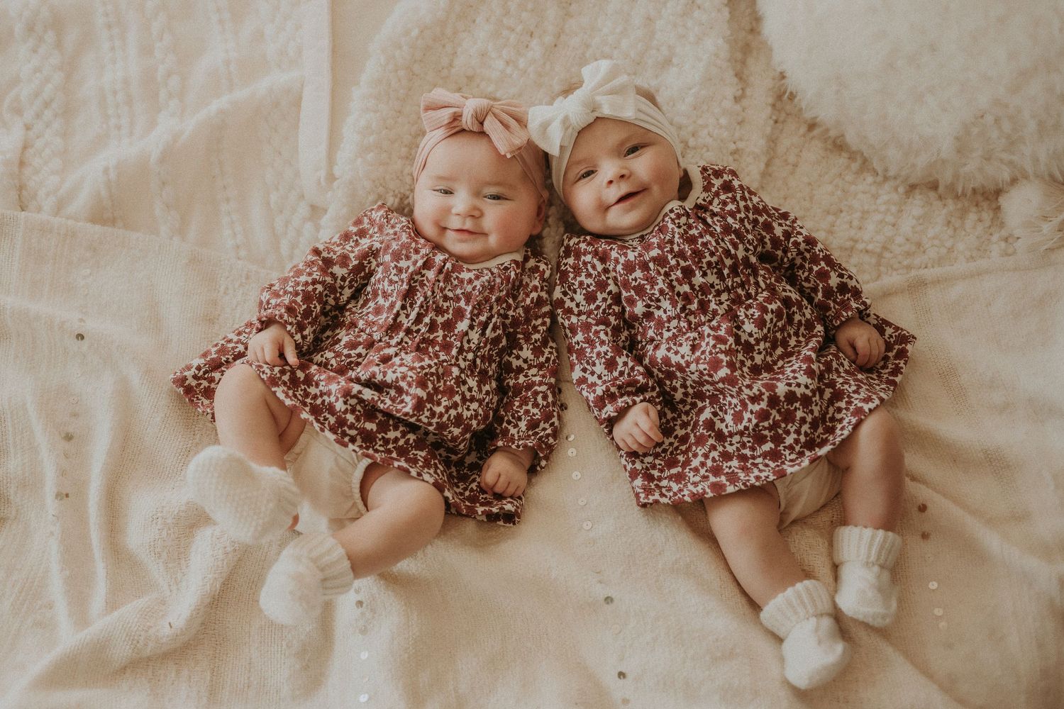 Twin babies wearing matching floral dresses lying on cream colored bedding.