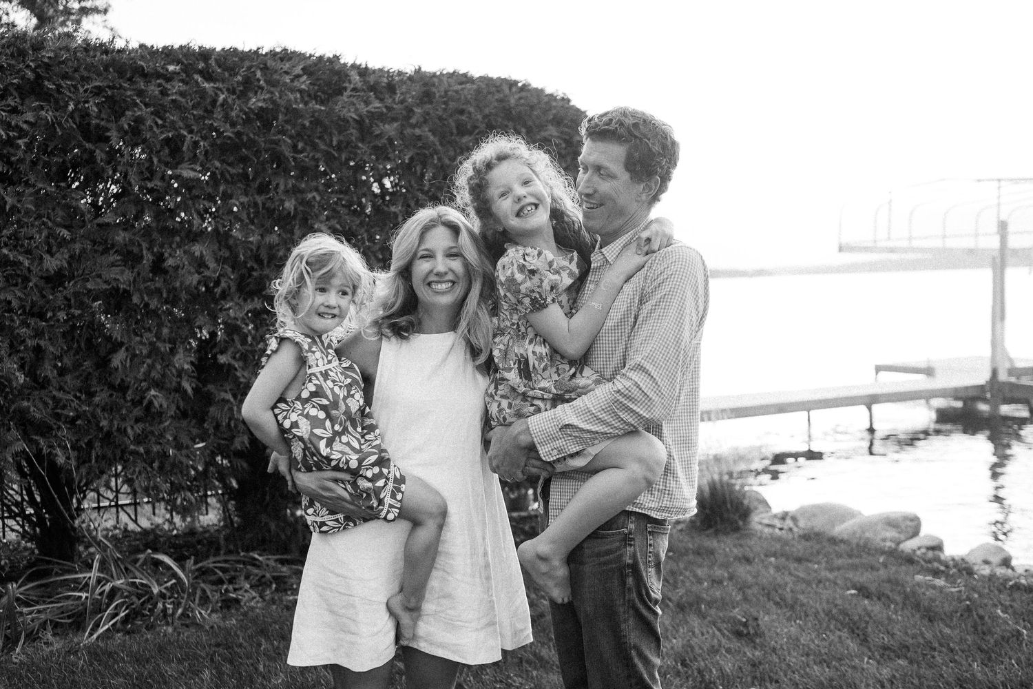 A family stands together outdoors in black and white, smiling by a waterfront with hedges in the background.