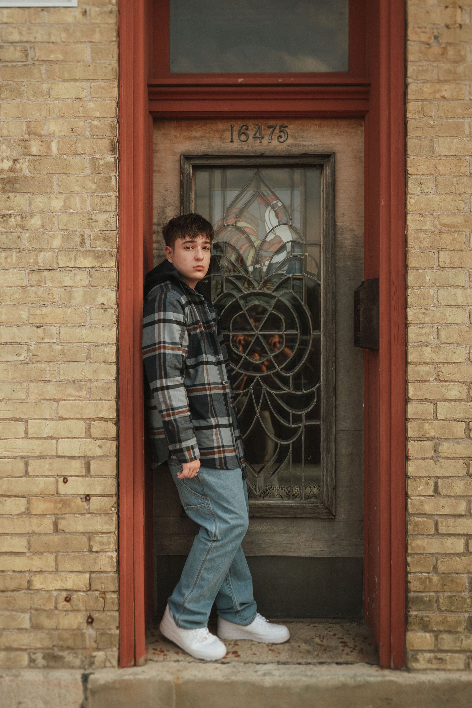 Person in plaid jacket and jeans posing in front of a vintage brick building with decorative windows.