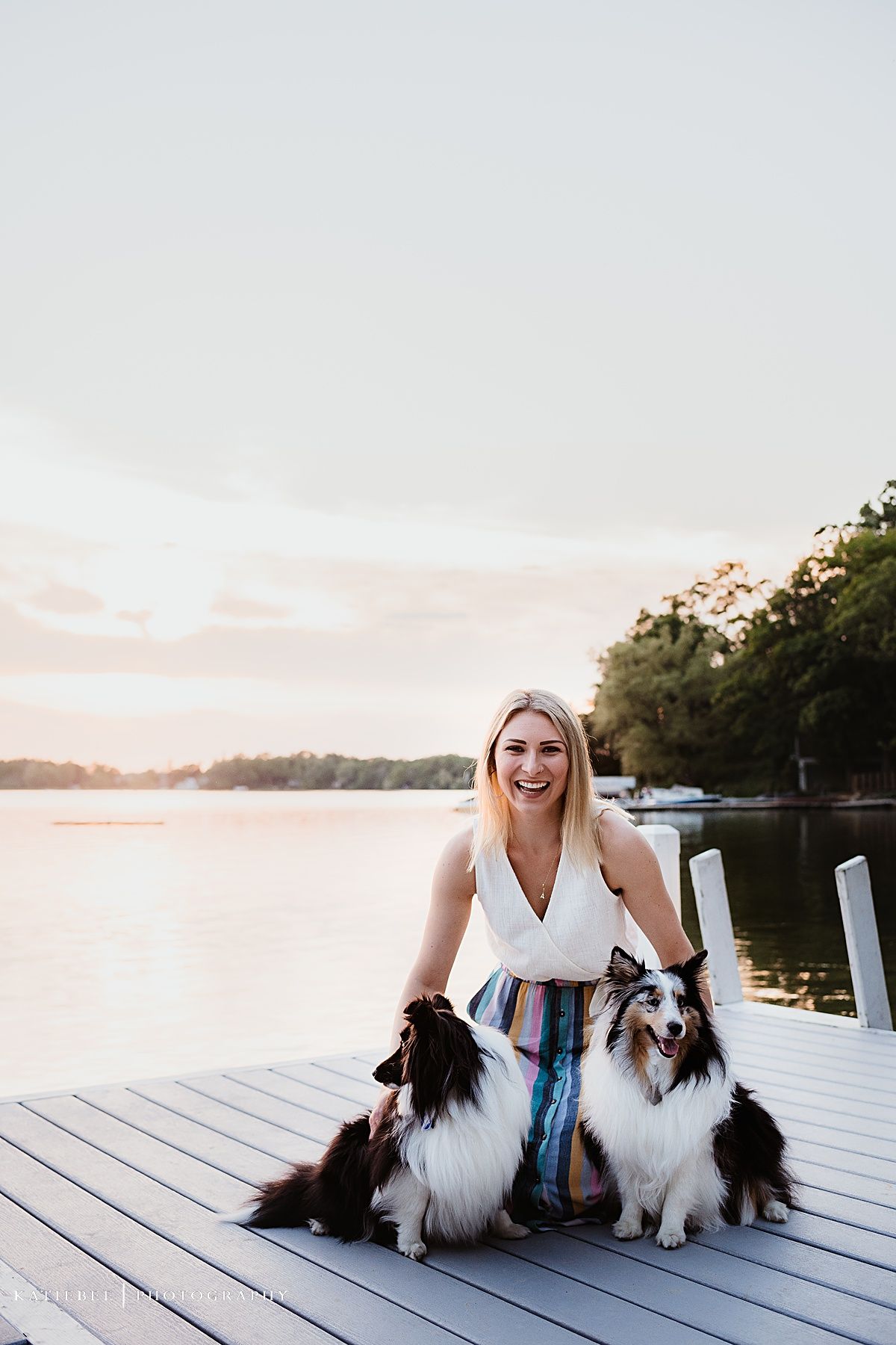 A woman sitting on a dock with two Sheltie dogs during sunset.