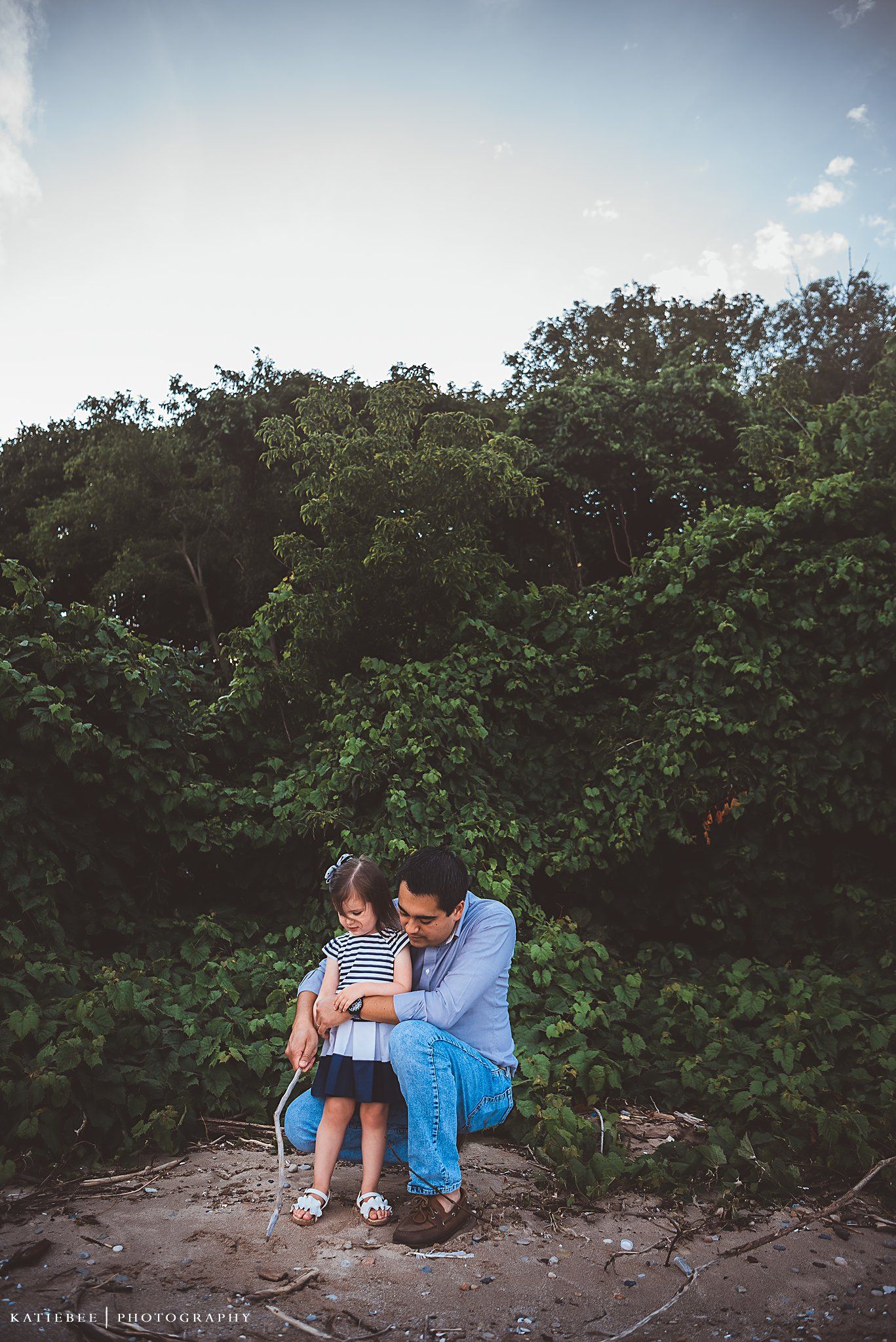 A touching outdoor portrait featuring a parent and child sitting together on rocky ground surrounded by greenery.