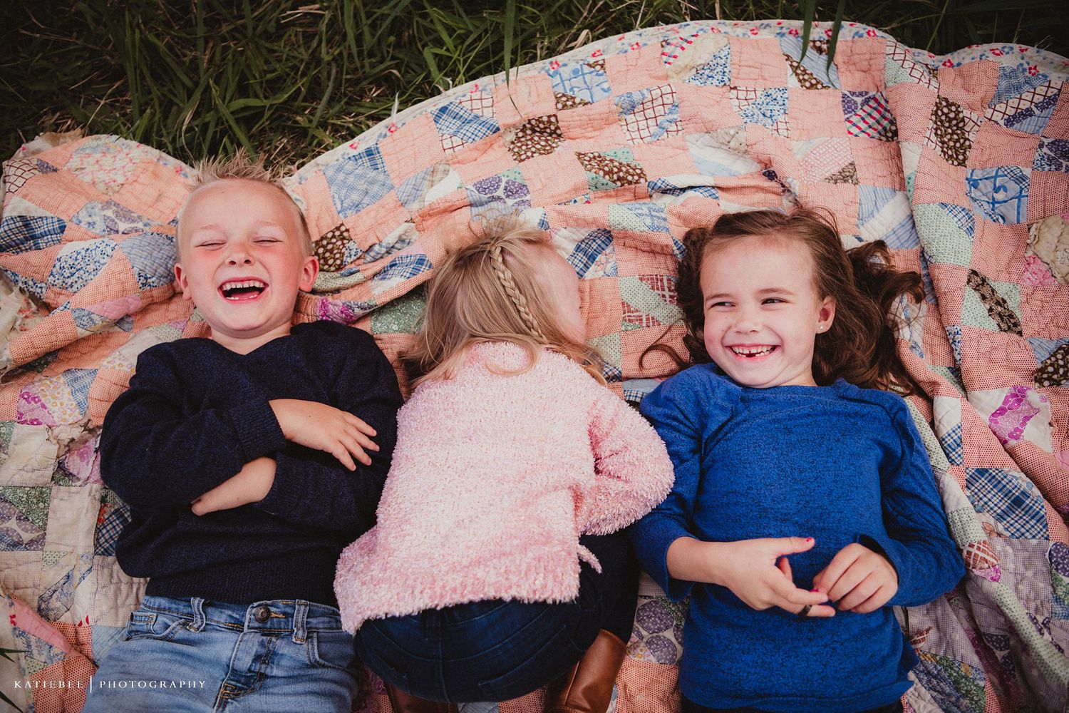 Three children laying on a floral blanket outdoors laughing together.