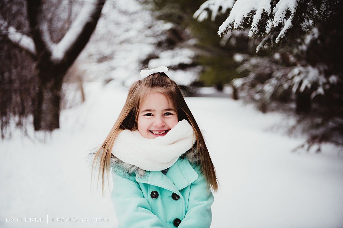 Young individual in mint green coat and white scarf smiling in snowy winter landscape.