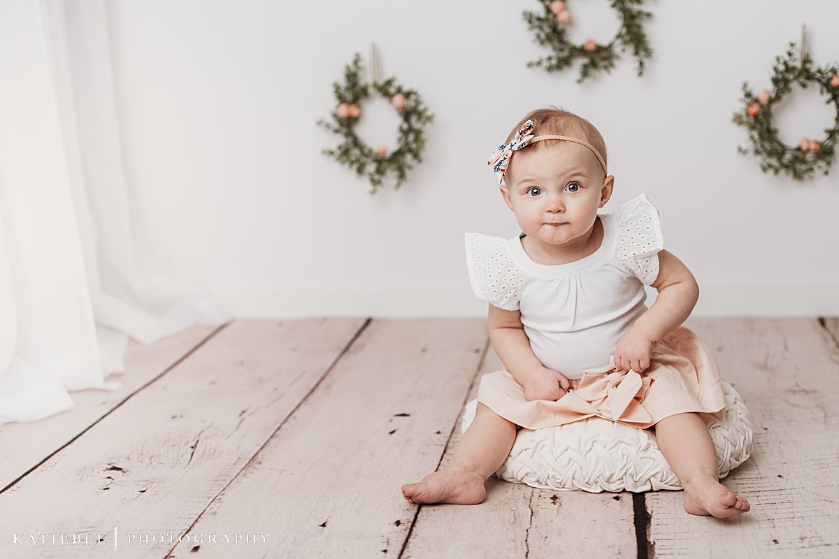 Baby in white dress sitting on wooden floor with wreaths decorating white wall background.