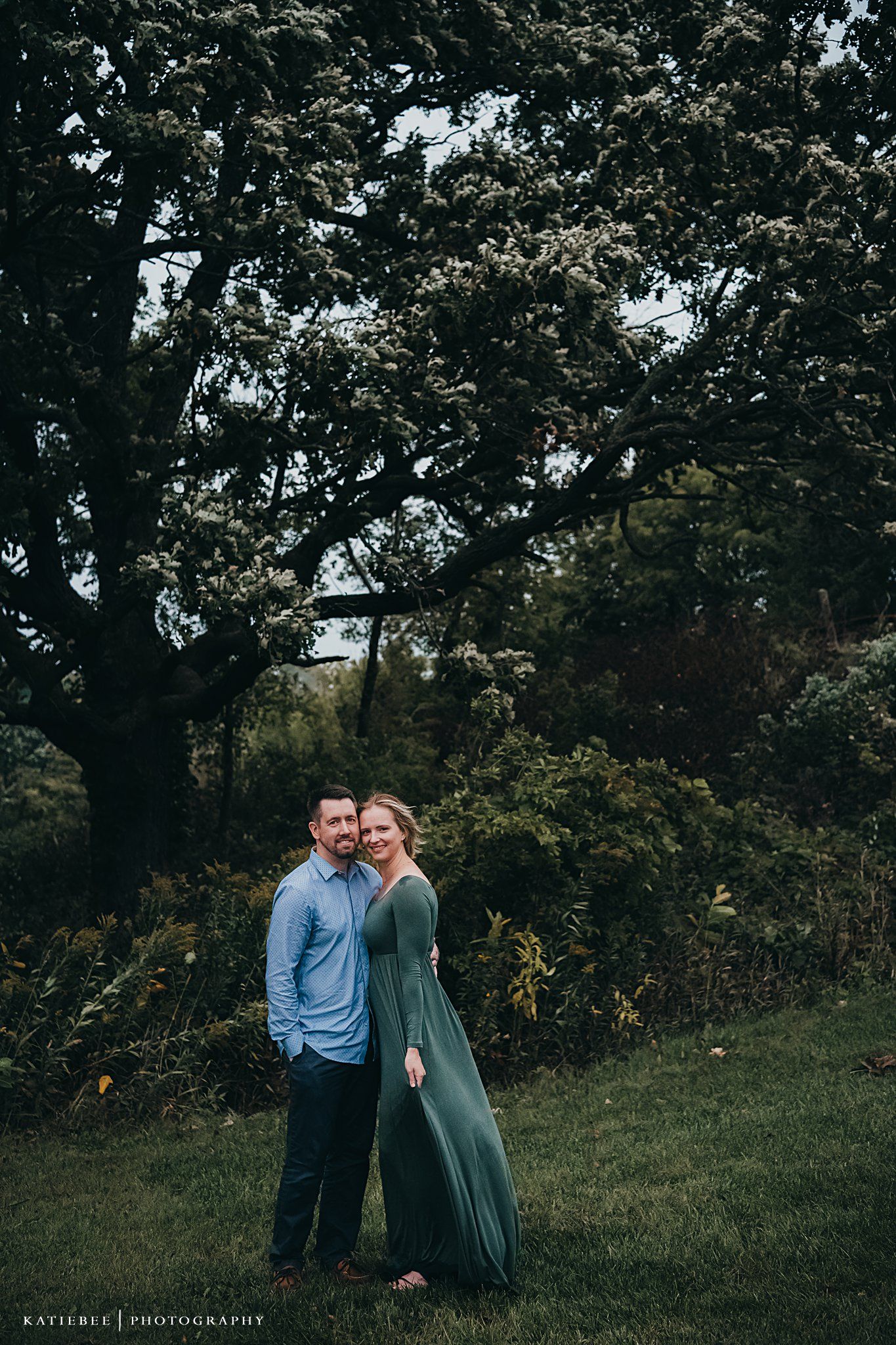 A romantic outdoor photo of a couple embracing under tall trees with the woman wearing a flowing sage green dress.