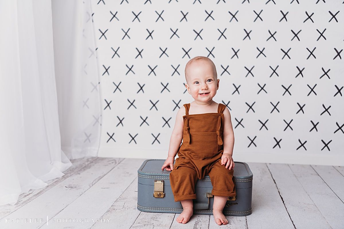 Baby in brown overalls sits on gray storage box against white patterned backdrop.