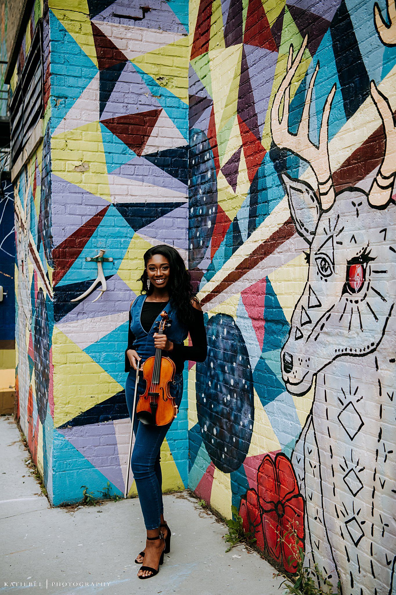 Musician with violin posing against colorful geometric street art with abstract animal design.