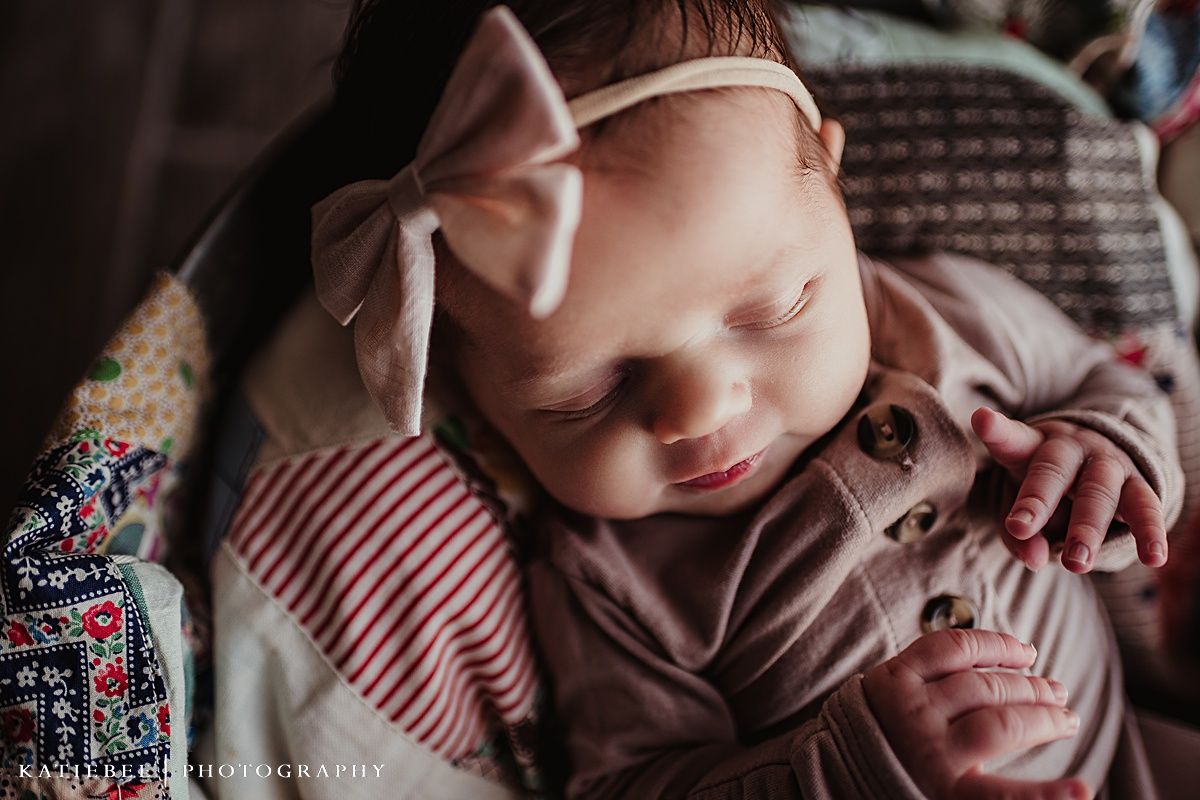 Sleeping infant wearing pink bow headband and beige outfit on patterned blanket.