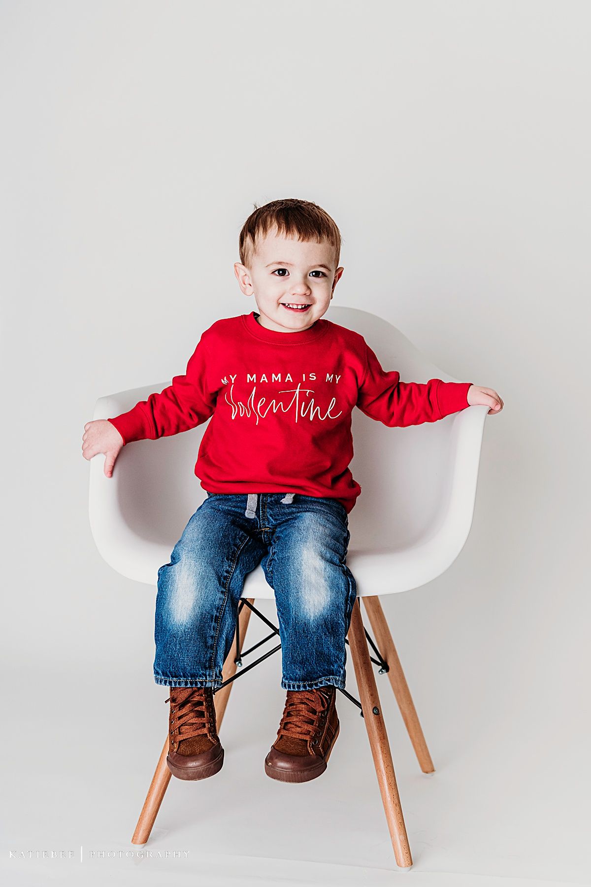 Small one sitting on white modern chair wearing red sweater with valentine message and brown boots.