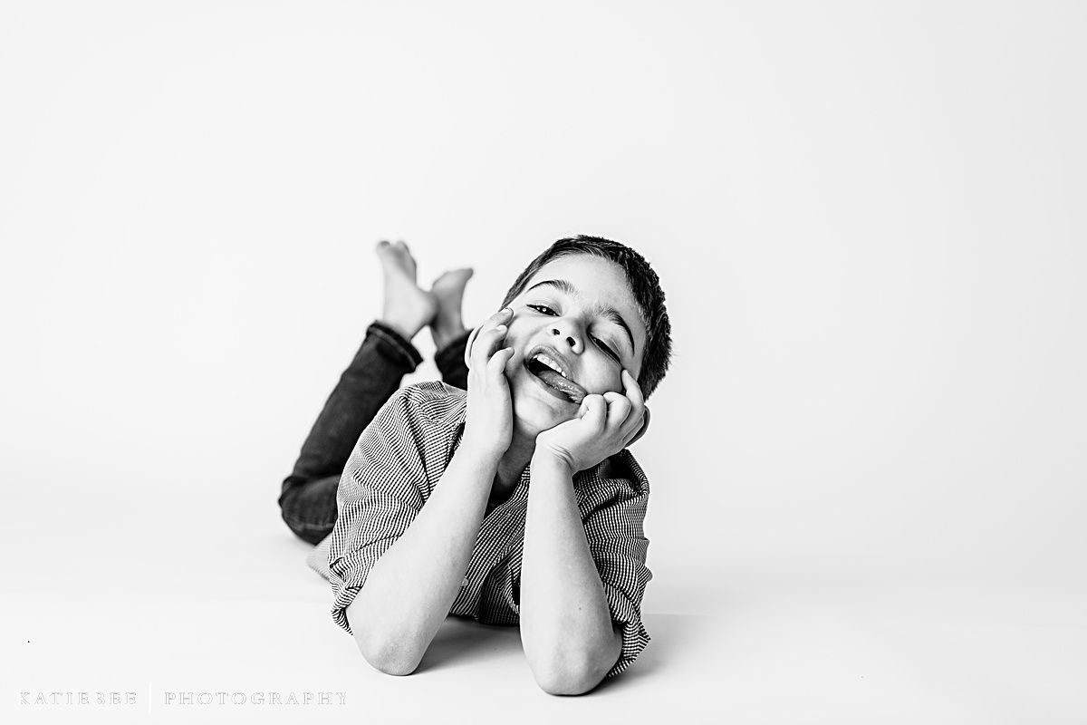 Child lying down with hands on face in playful black and white portrait.