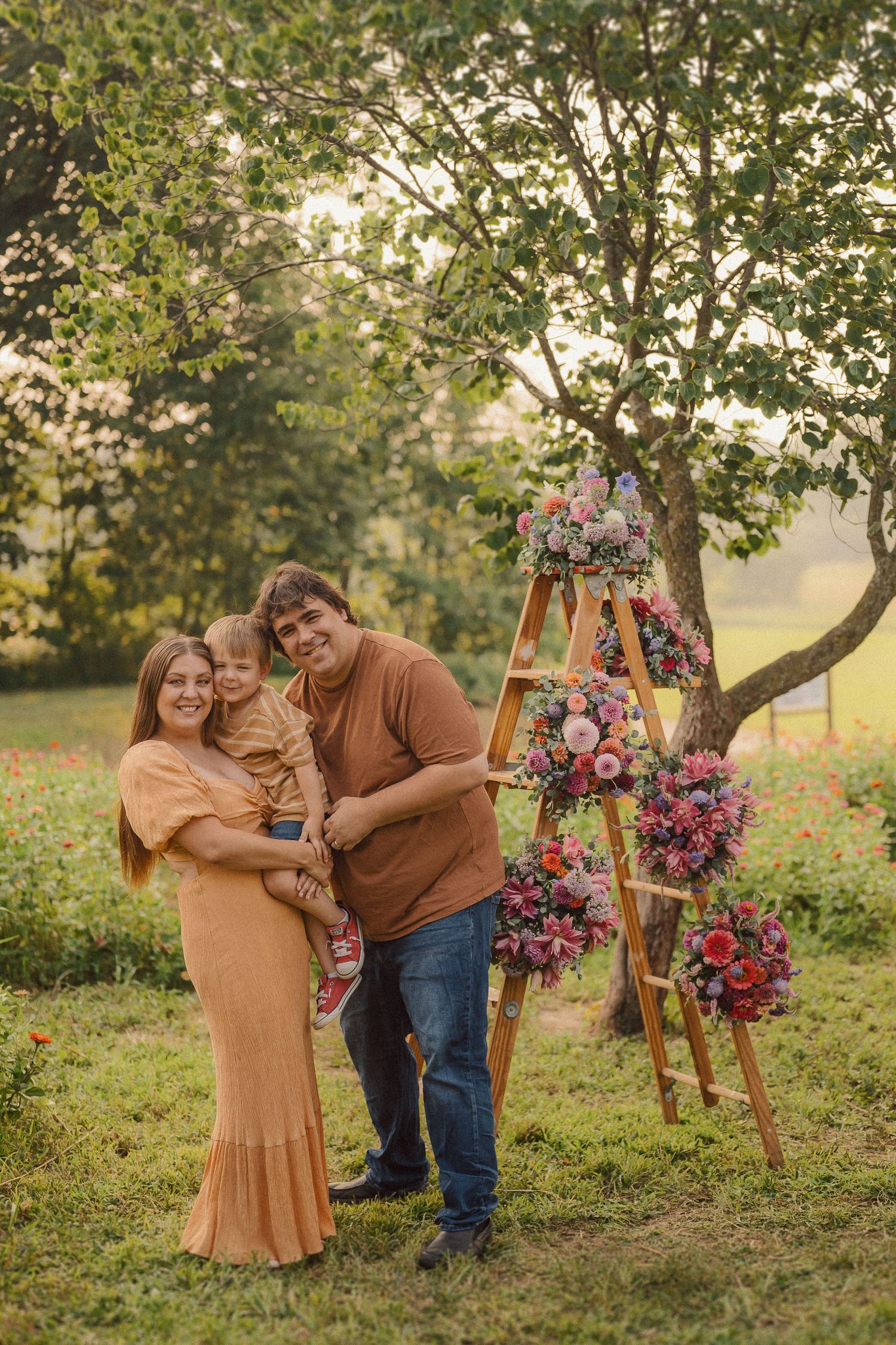 A couple in coordinating earth tones poses with their child by a rustic flower-adorned ladder in a garden setting.