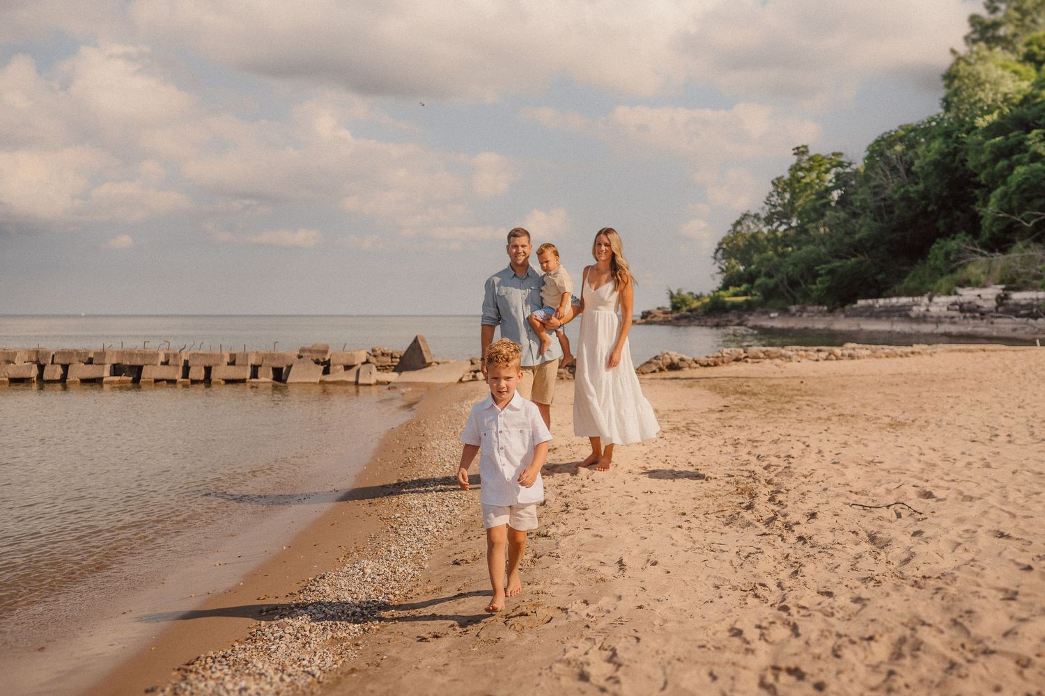 Family walking along peaceful shoreline with cloudy sky backdrop.