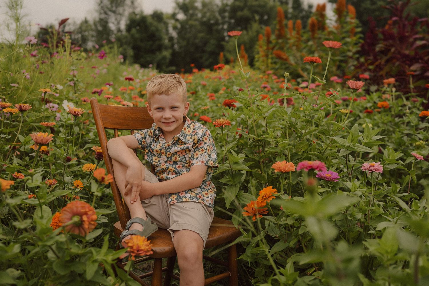 Young person sits among vibrant orange and pink zinnias in a lush summer garden.