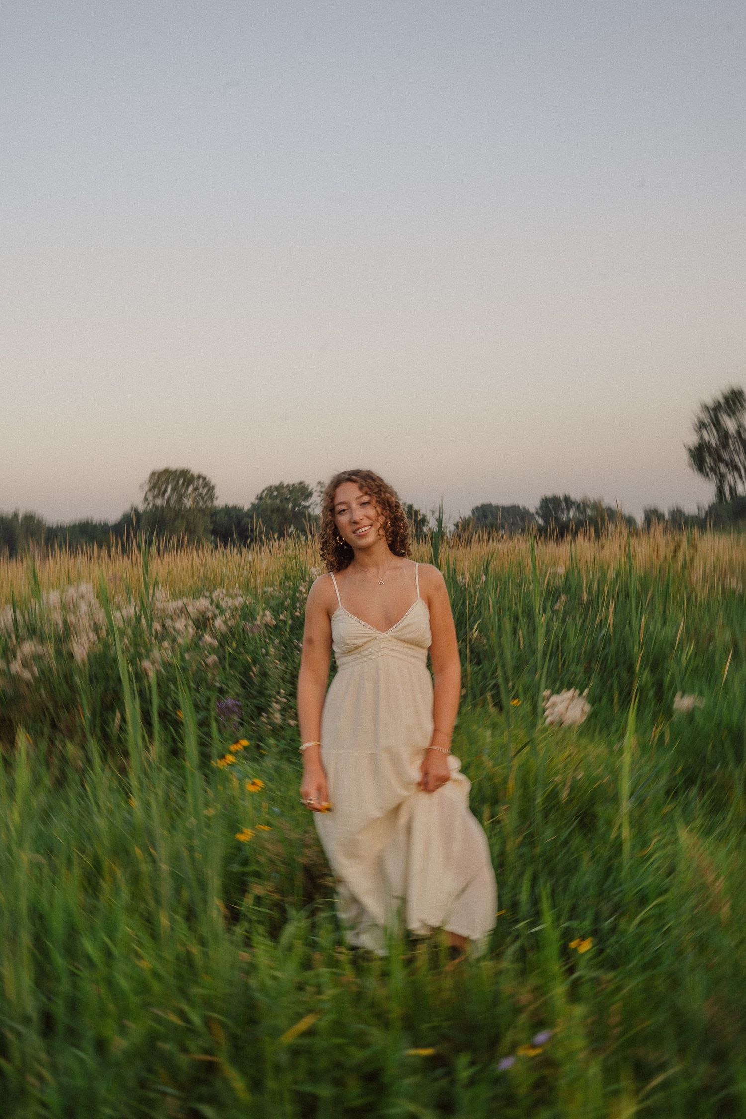 A person in a flowing cream dress stands among tall wild grasses during sunset.