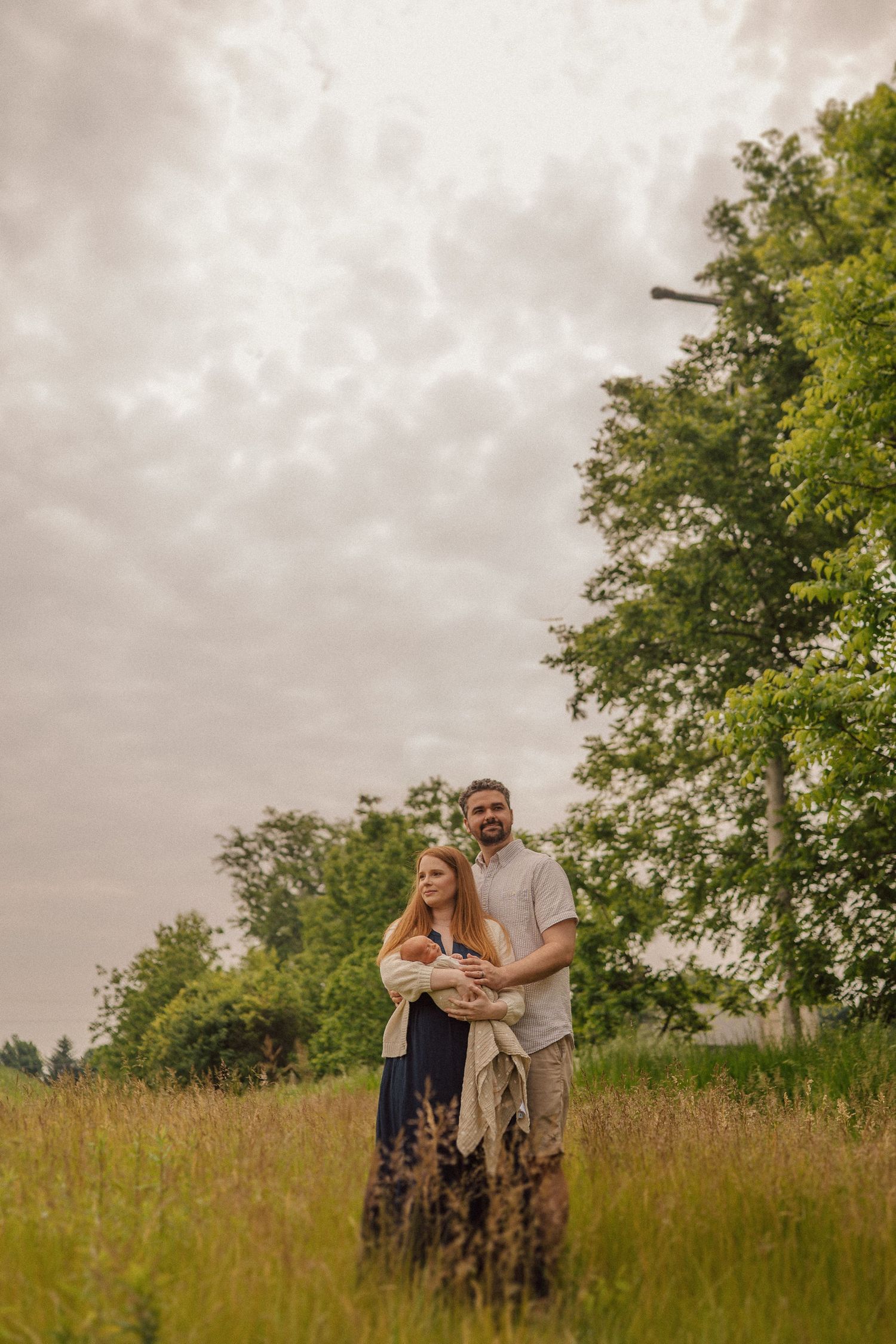 Couple embracing in tall grass field under dramatic cloudy sky with trees in background.