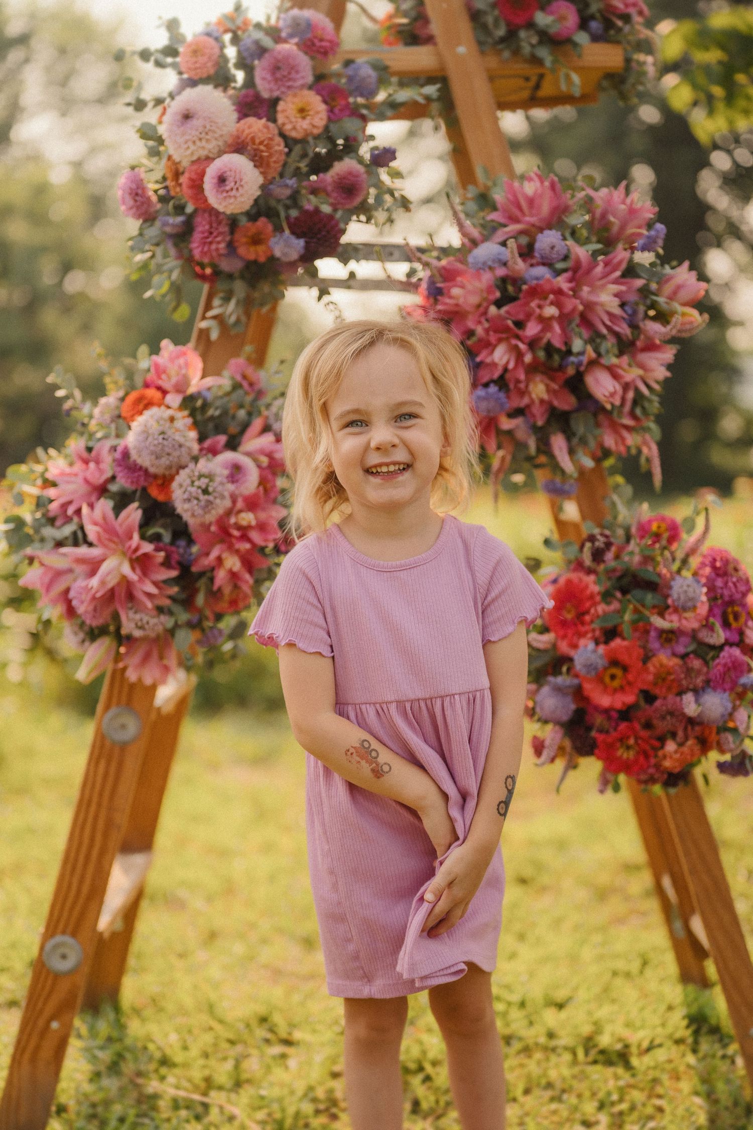 Wooden triangle arch decorated with pink and purple floral arrangements on a sunny garden lawn.