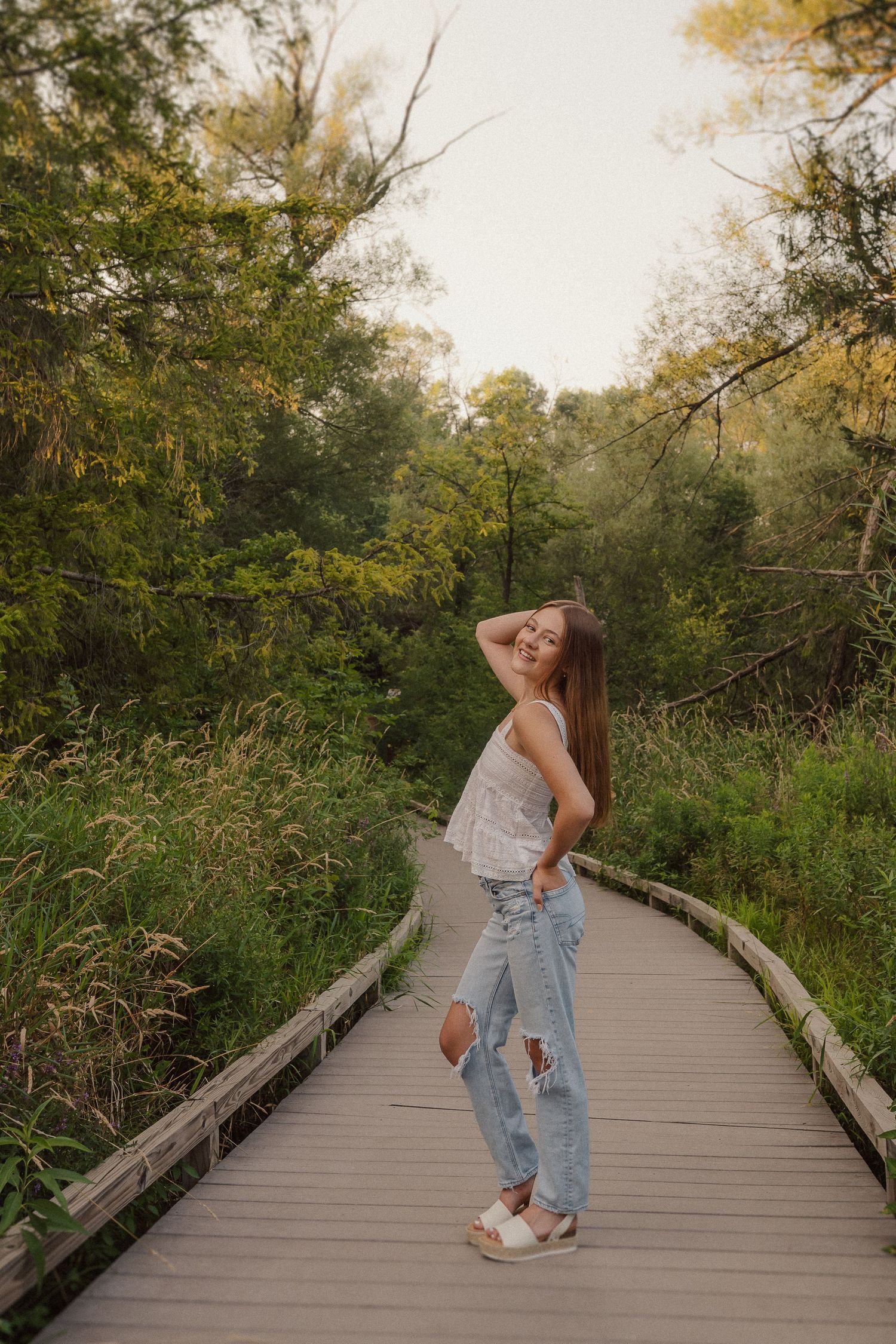 A person in denim overalls poses in a series of dance-like movements on a wooden boardwalk surrounded by lush greenery.