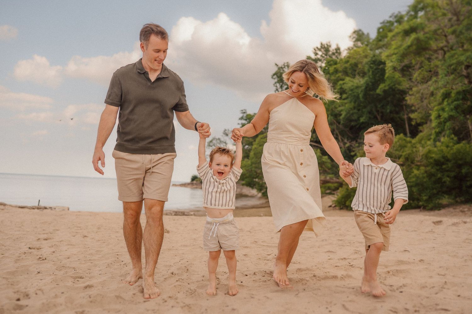 A family holds hands and walks together along a sandy beach at sunset, wearing coordinated beige and white outfits.