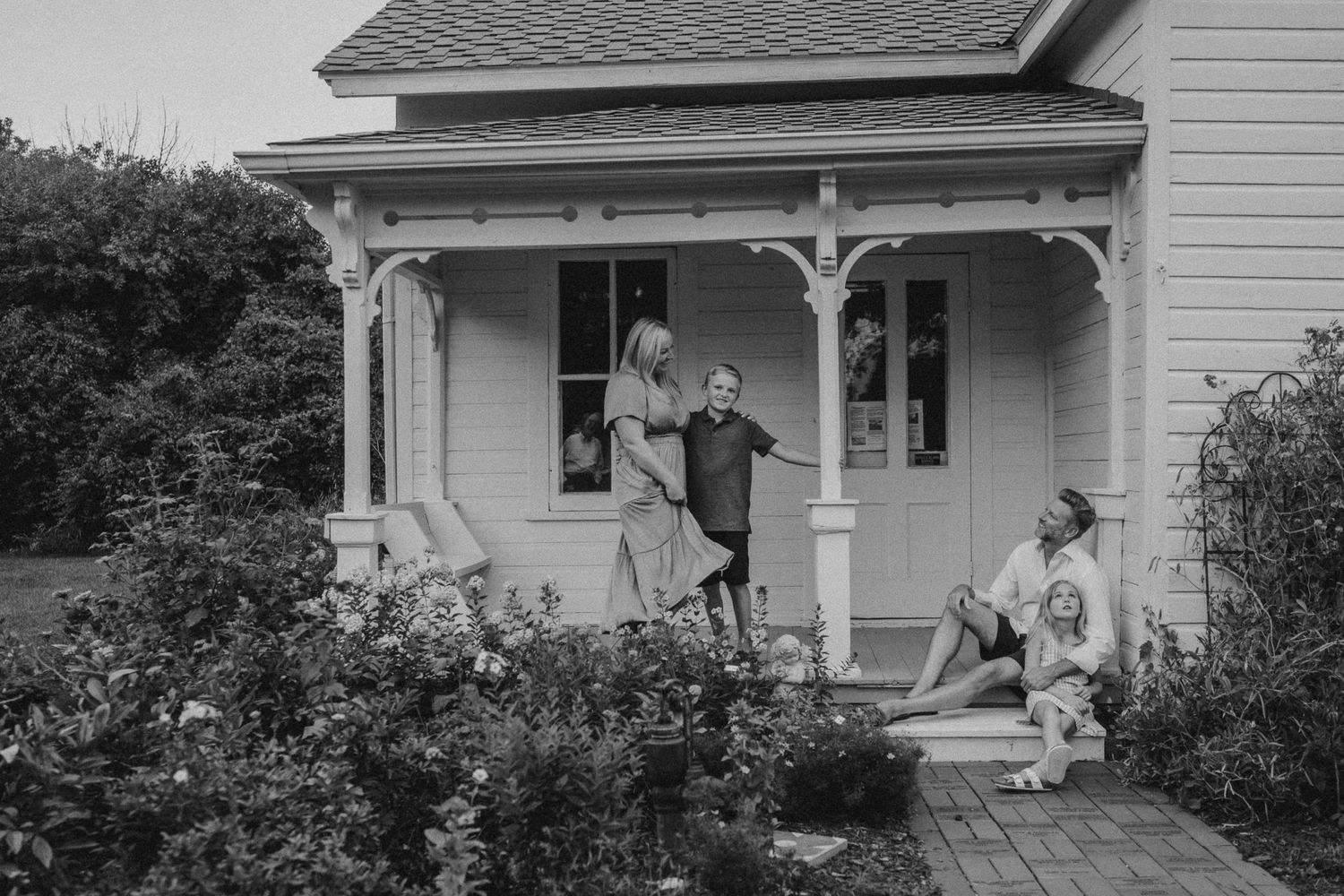 Black and white image of family gathered on white farmhouse porch surrounded by garden.
