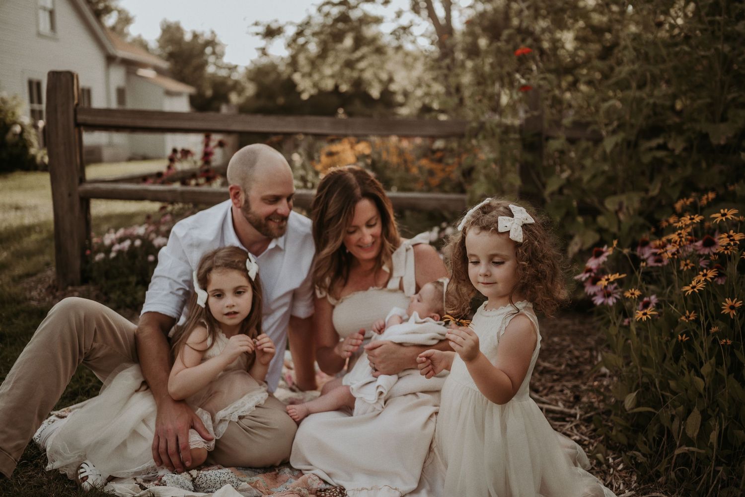 Family in coordinated white and neutral outfits sits together on blanket in garden setting.