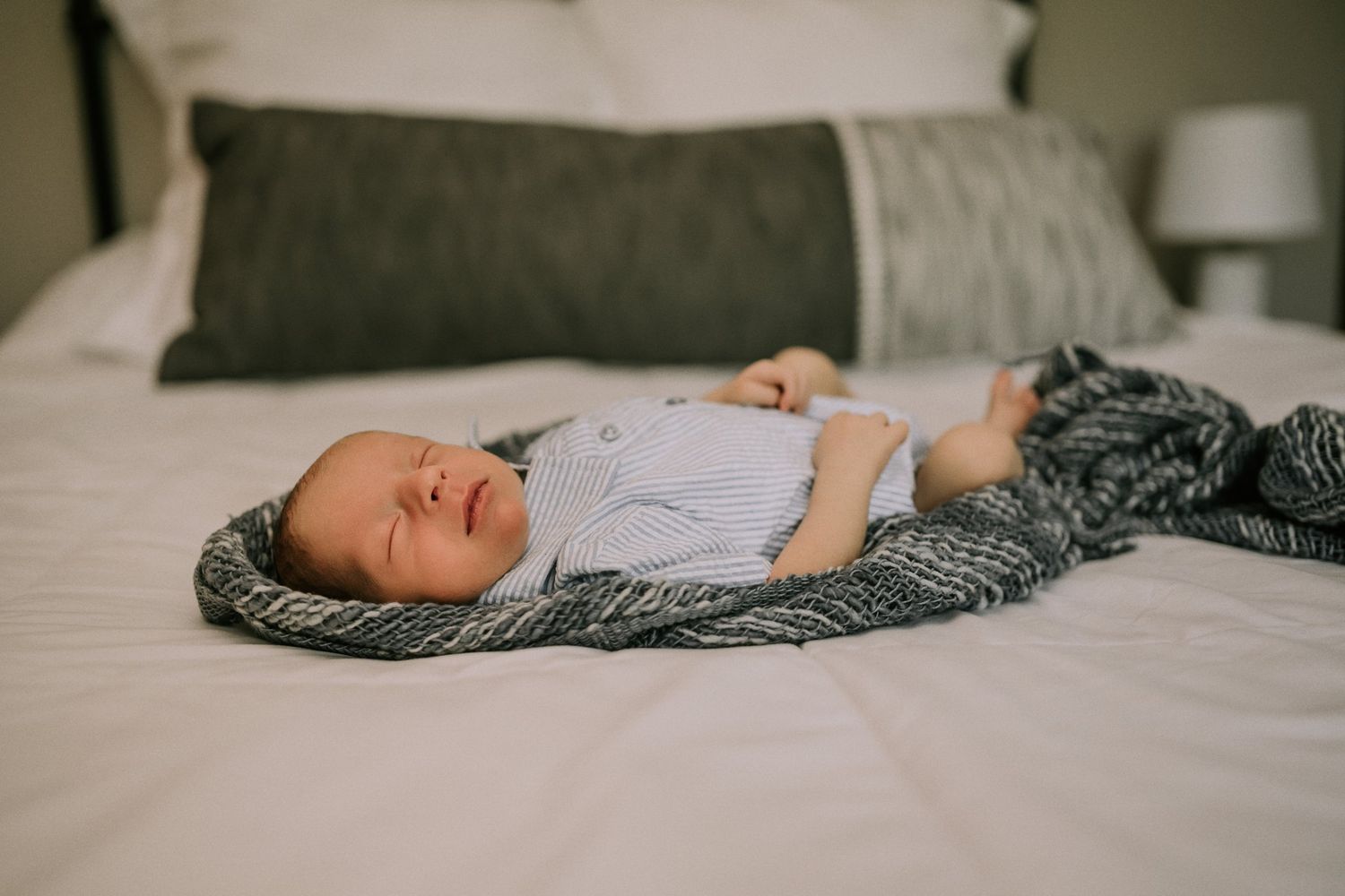 Sleeping newborn rests peacefully on gray knit blanket on white bedding.