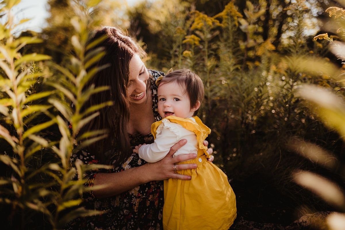 A tender outdoor portrait of parent and baby in a yellow dress surrounded by golden sunlit foliage.