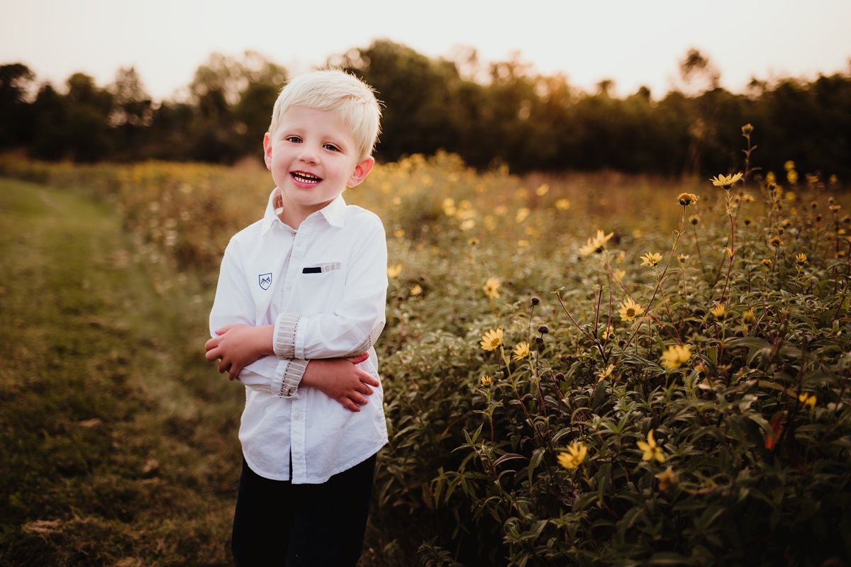 Child in white button-down shirt stands confidently in field of yellow wildflowers at sunset.
