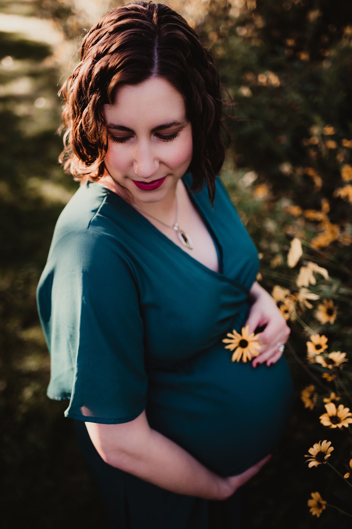 Expectant mother in teal dress poses with yellow flowers during sunset maternity photoshoot.