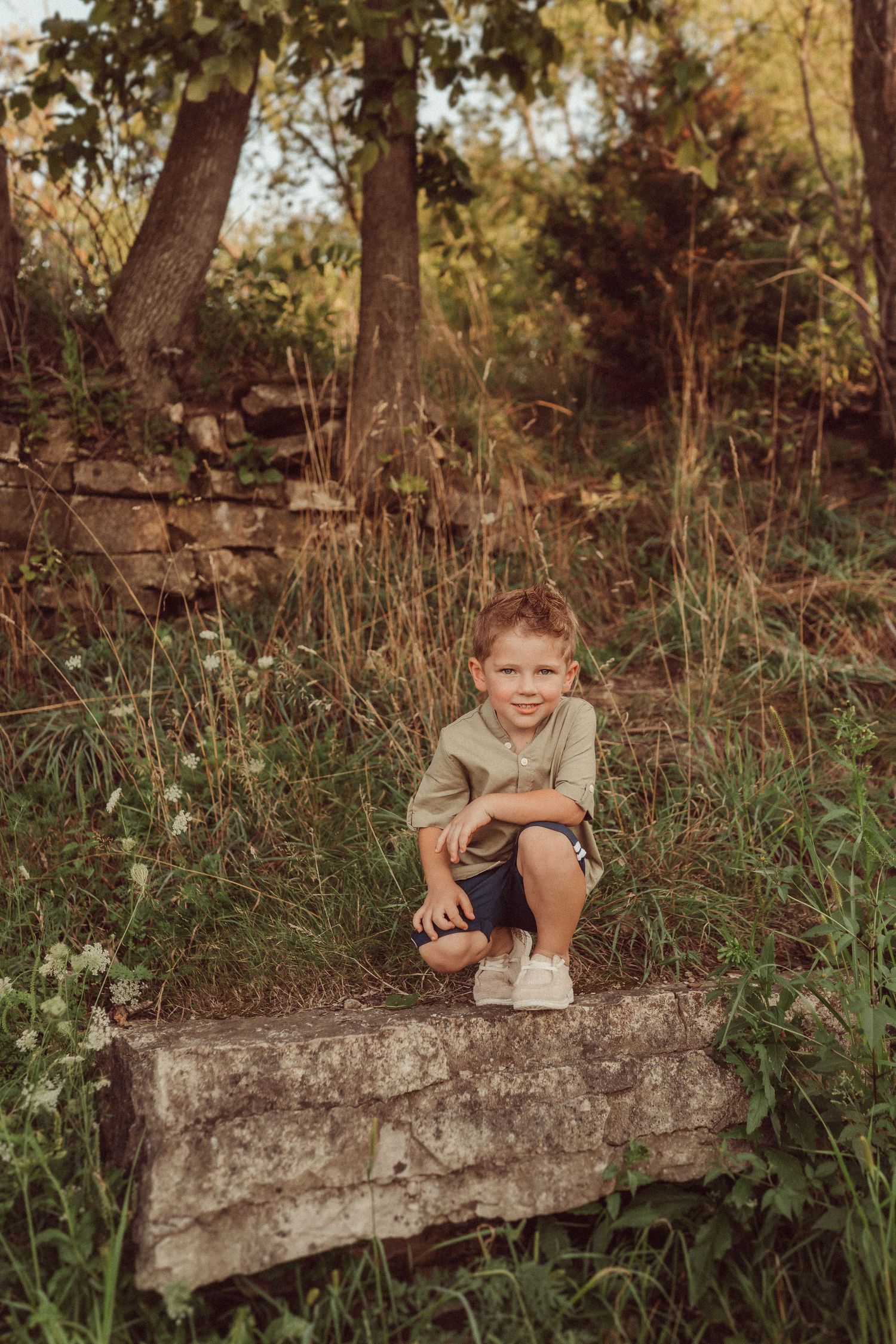 Child crouches on stone ledge in rustic woodland setting surrounded by tall grass.