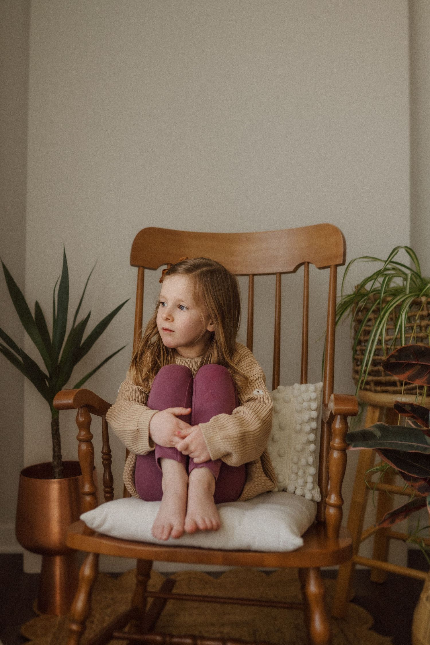 Thoughtful pose in wooden rocking chair with purple leggings and cozy beige sweater.
