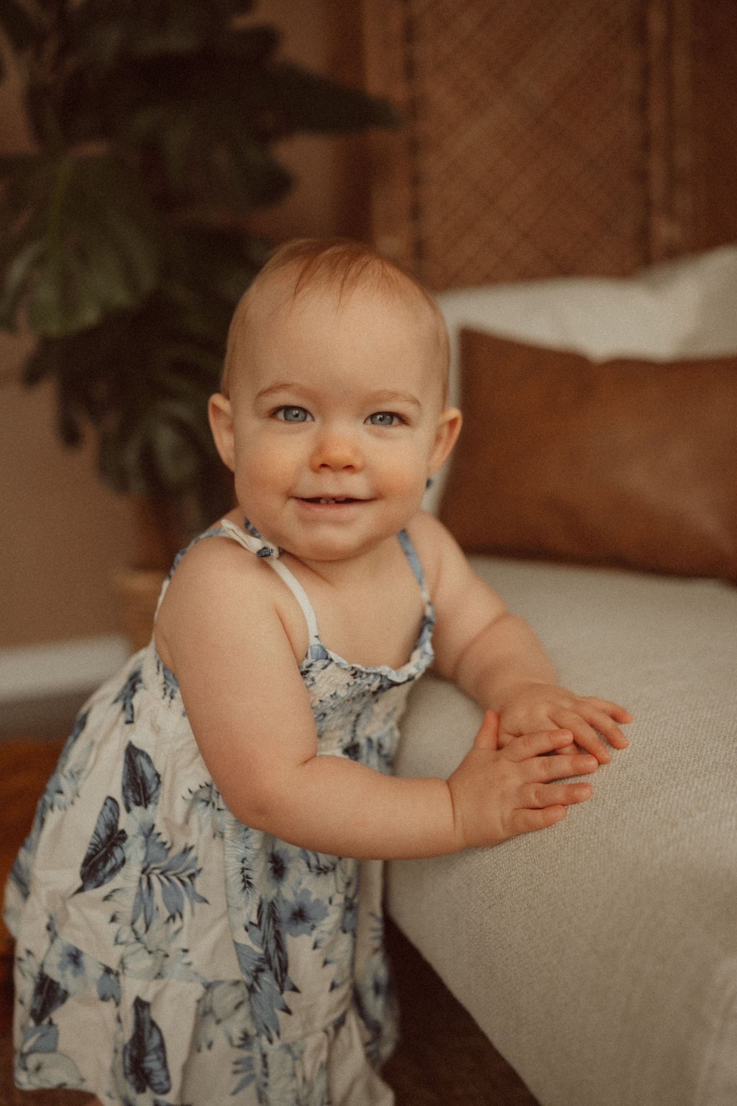 A small child in a floral dress kneels next to a light-colored couch.