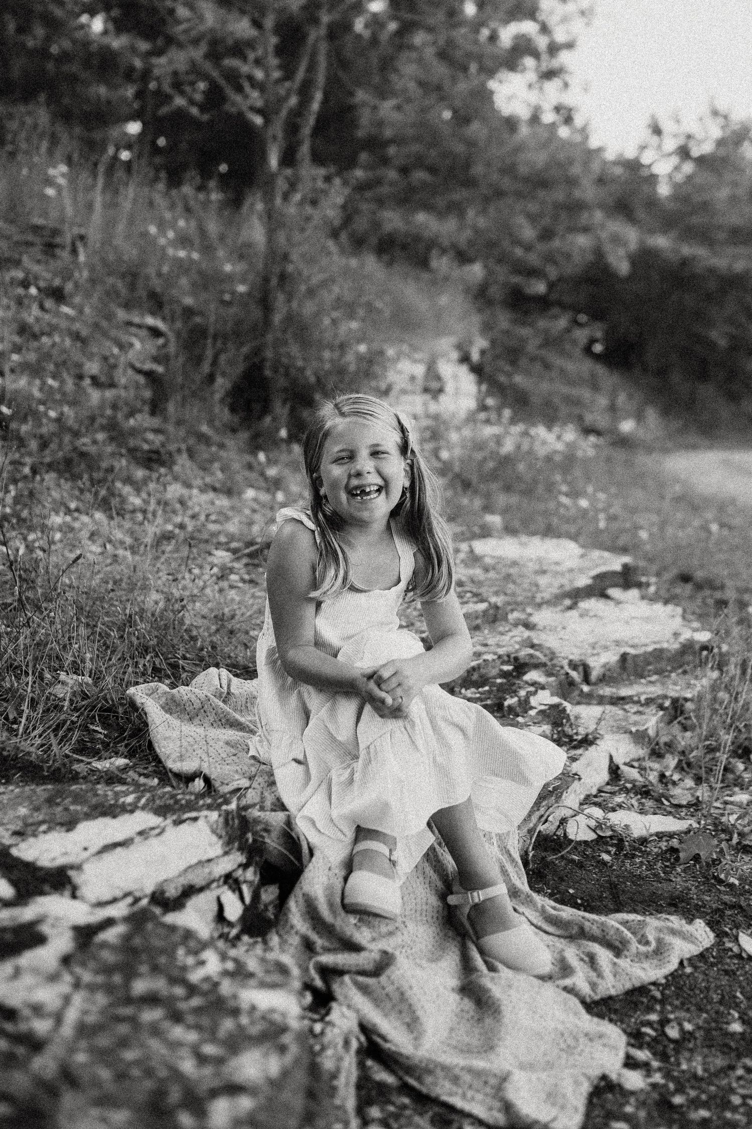 A black and white portrait of a child laughing while sitting on a blanket outdoors.