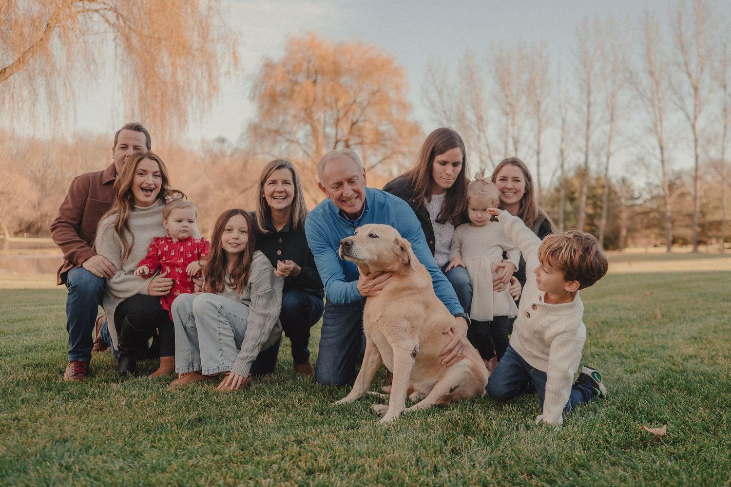 A group sits together on grass with a golden retriever dog during an autumn photoshoot with bare trees in background.