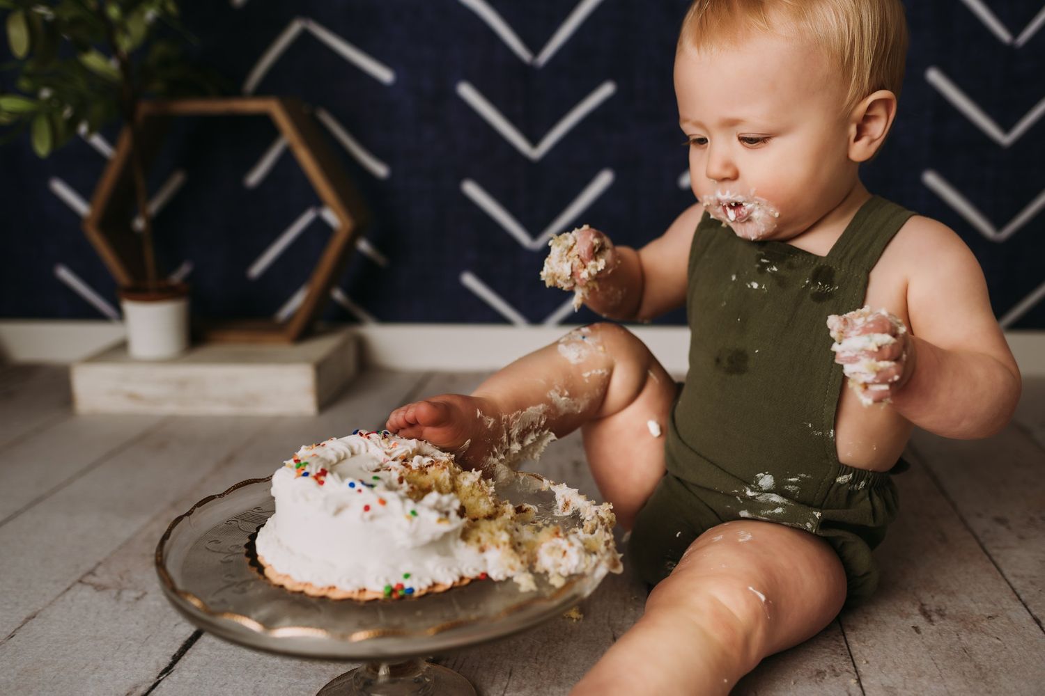 A joyful cake smash scene with scattered frosting on a glass pedestal against geometric wall art.