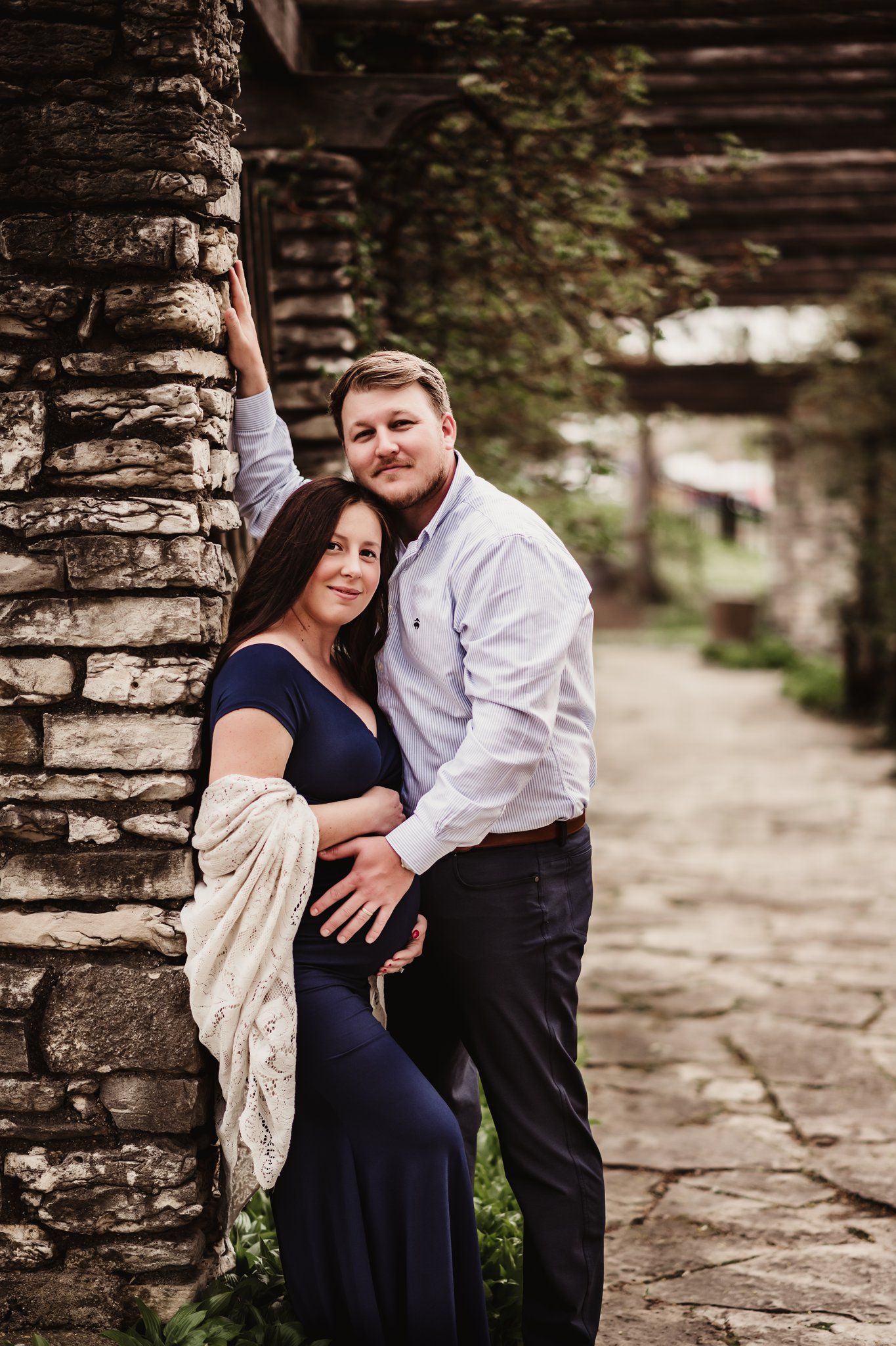 Maternity portrait showing couple embracing by stone pillar under garden archway.