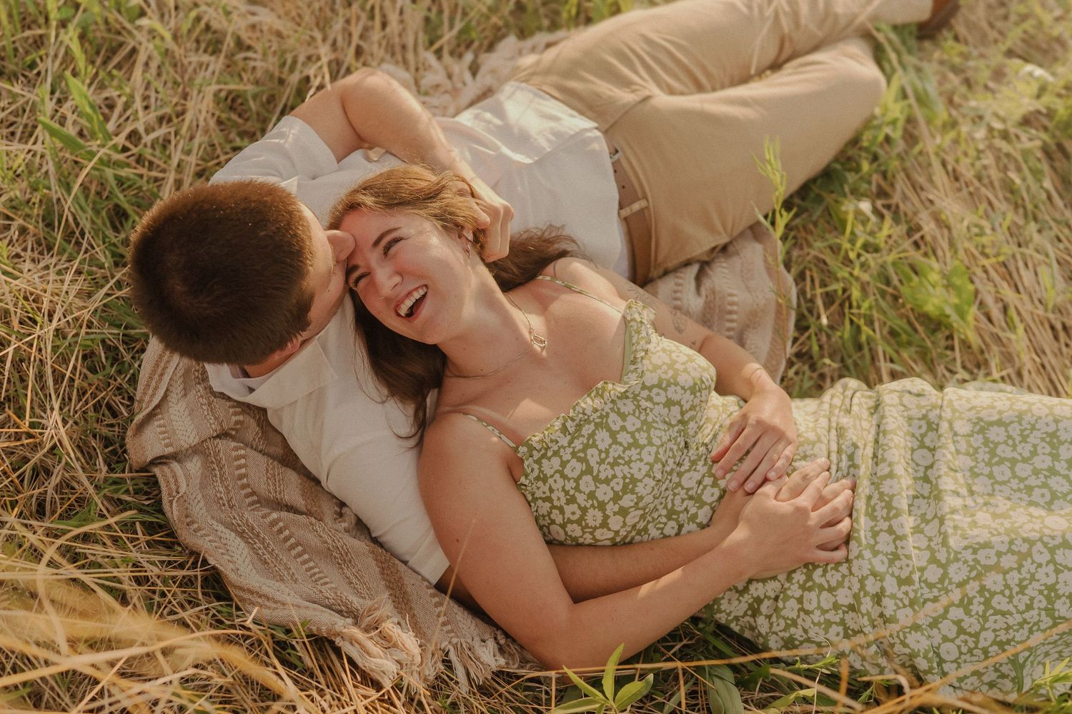 A romantic couple shares a joyful moment while laying in a grassy field at sunset.