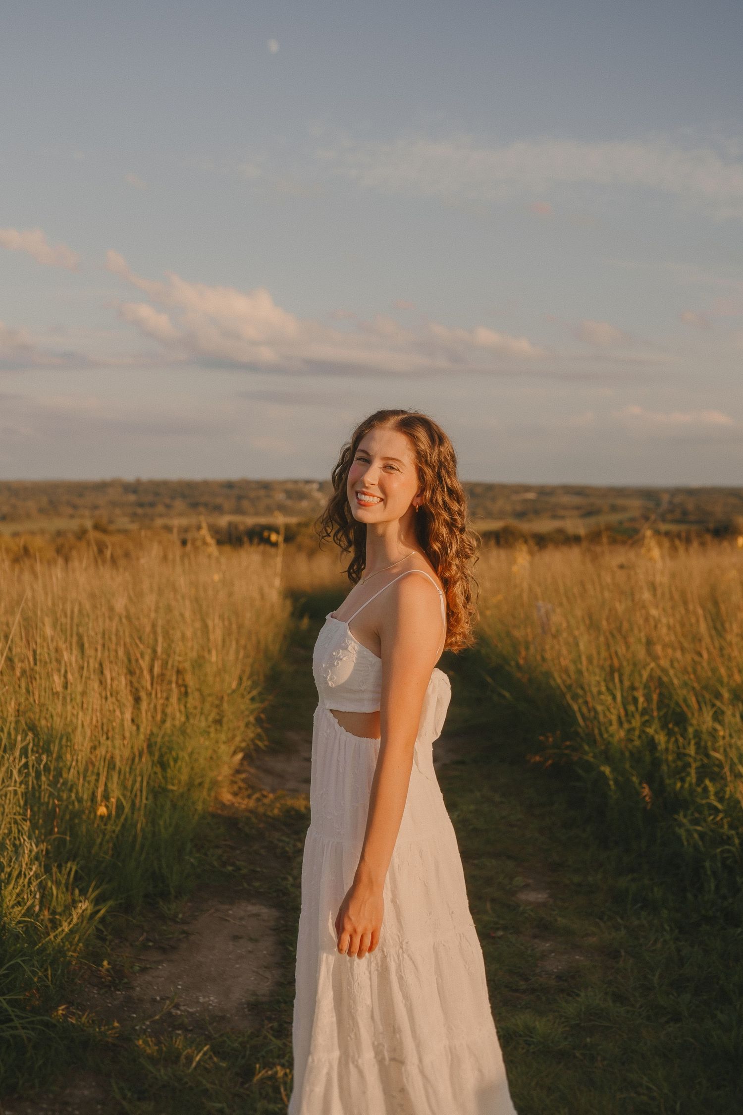 A person in a white summer dress smiles while standing on a dirt path amidst golden prairie grass.