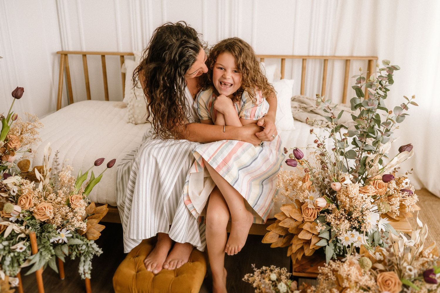 Tender moment captured on white bed with floral arrangements and natural light streaming through window.