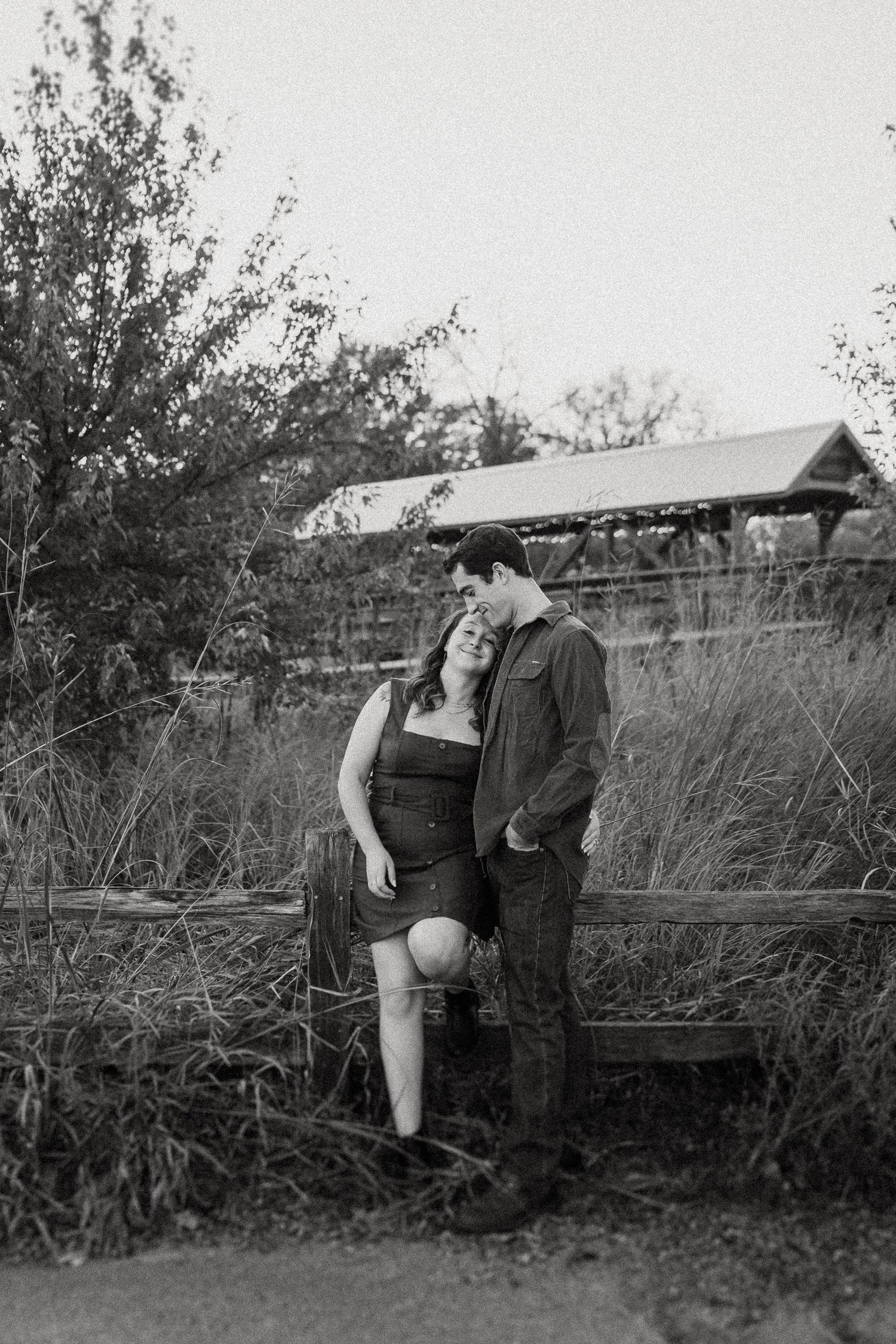 A romantic couple embraces beside a rustic wooden fence in a black and white photograph.