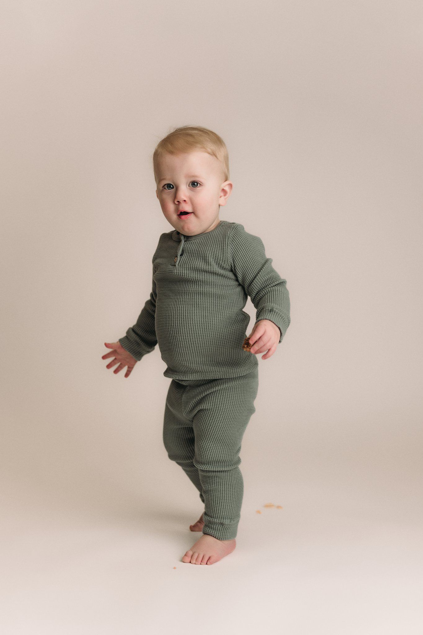 Full body shot of toddler standing in sage green ribbed outfit against neutral background.