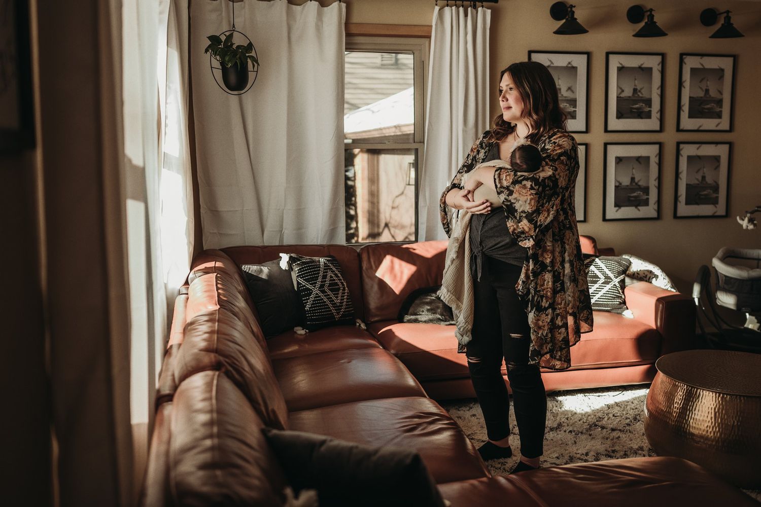 A pregnant woman in a floral dress stands by a window in a cozy living room.