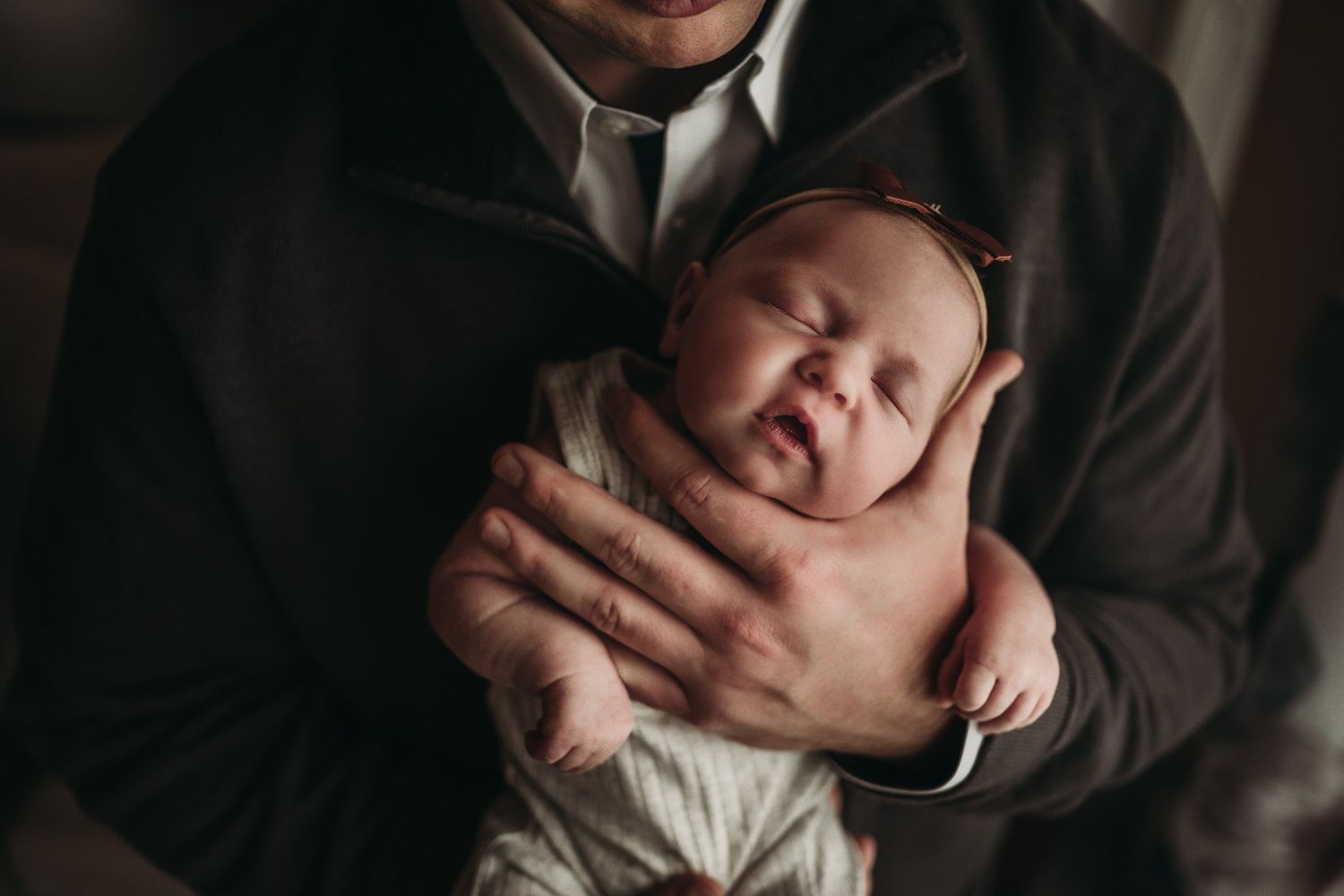 A parent in dark clothing tenderly holds their alert newborn baby near a window.
